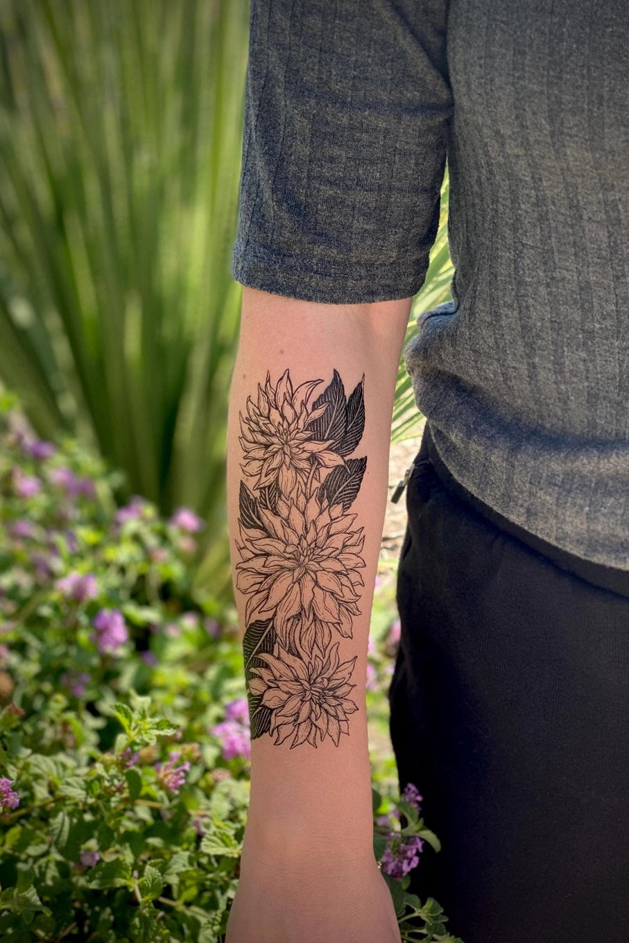 Person wearing a gray shirt with a floral tattoo on their arm, standing in a garden. ©Nature Tats