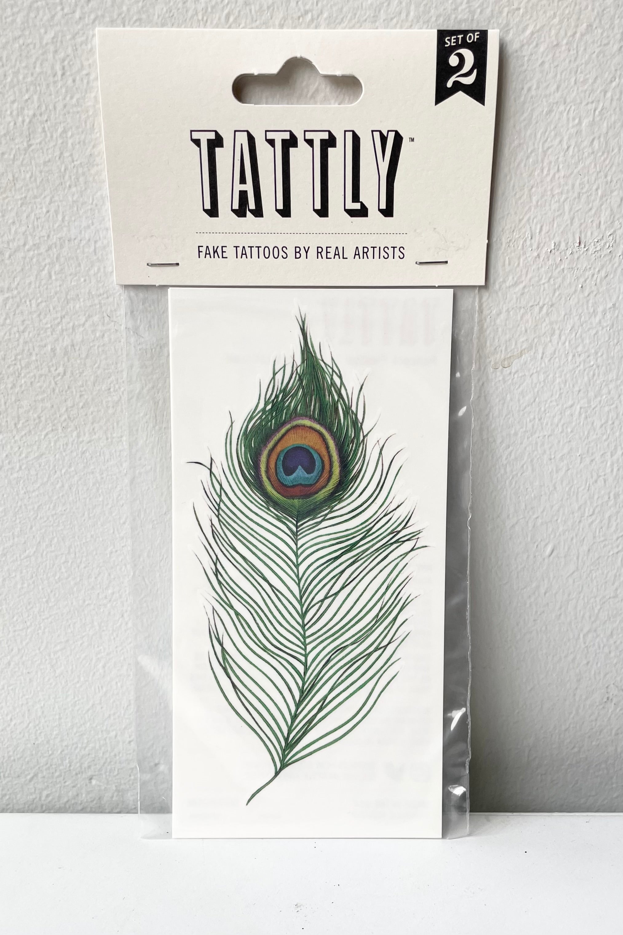 The peacock feathers tattoo in its Tattly packaging against a white wall.