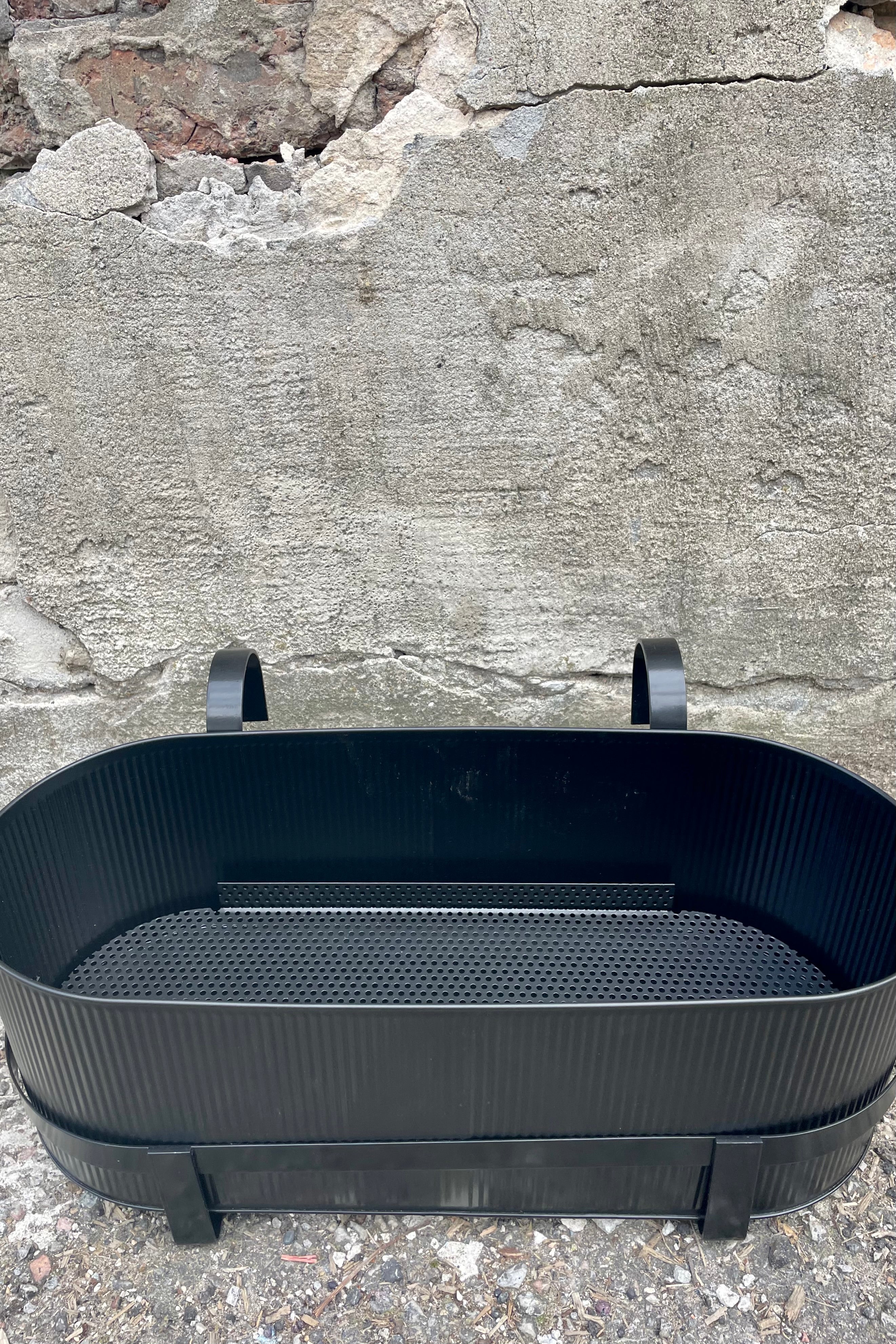 Photo of Ferm Living's Bau Balcony Box in black. The planter features a horizontal holder and bracket and is oval in overall shape. There is a vertical texture to the metal planter. Photo is taken showing the sides and top / inside of the planter. It is photographed on the ground against a cement wall.