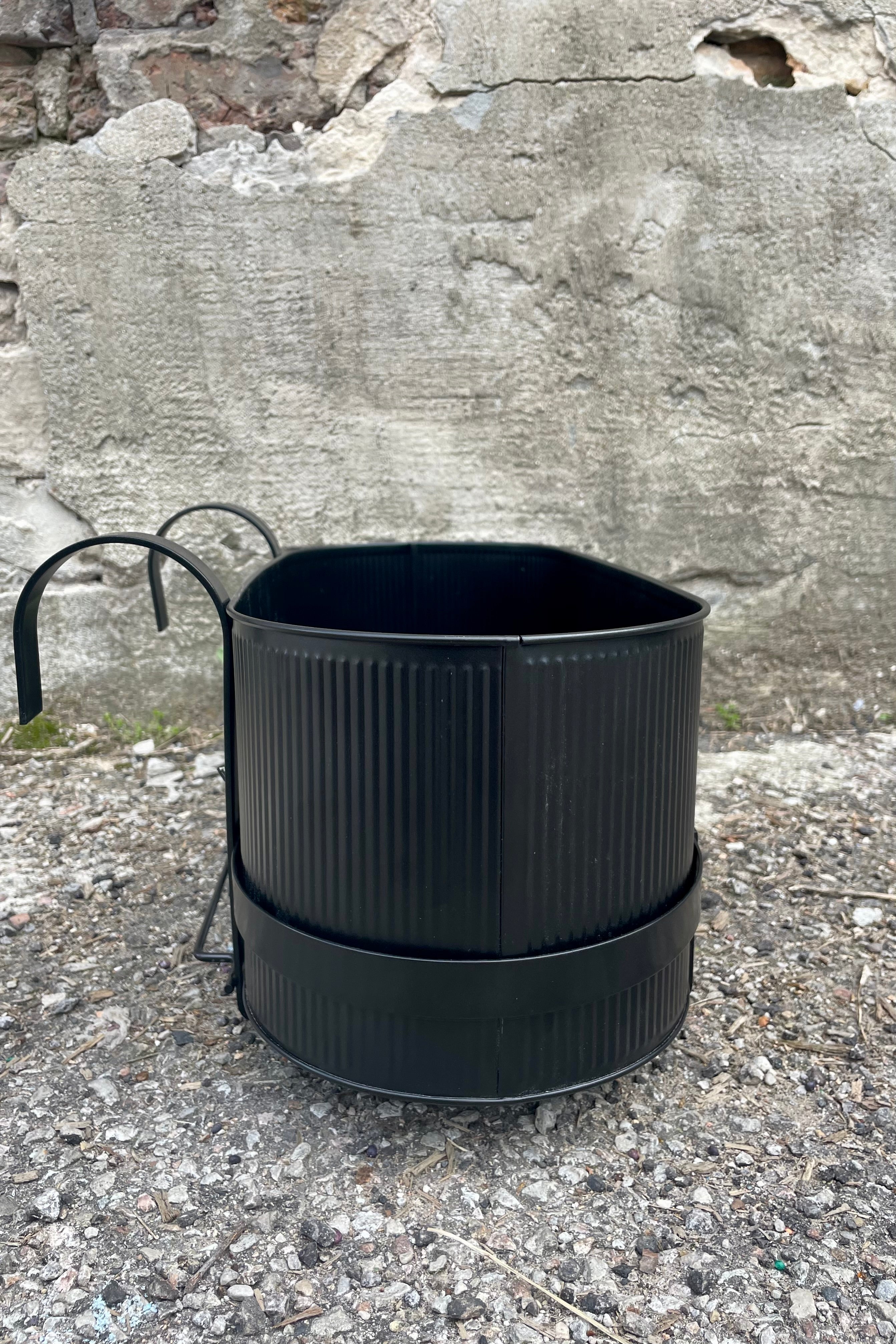 Photo of Ferm Living's Bau Balcony Box in black. The planter features a horizontal holder and bracket and is oval in overall shape. There is a vertical texture to the metal planter. Photo is taken showing the sides and top / inside of the planter. It is photographed on the ground against a cement wall. The view is from the size showing the narrow shape of the planter.