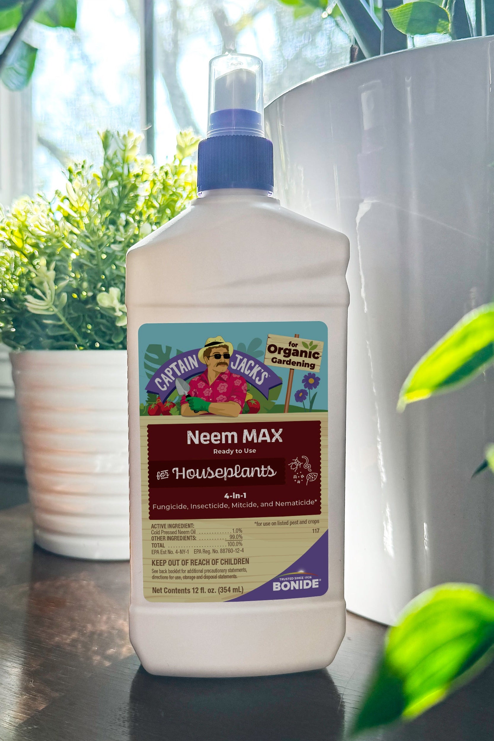 A bottle of Bonide Captain Jacks Neem Max Neem Plant & Houseplant liquid fertilizer standing on a dark wooden table, surrounded by potted green plants in a bright indoor setting. ©Bonide