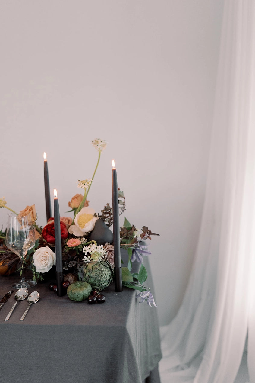 Mo & Co Charcoal beeswax taper candles shown lit and in a table setting with fruit and flowers.