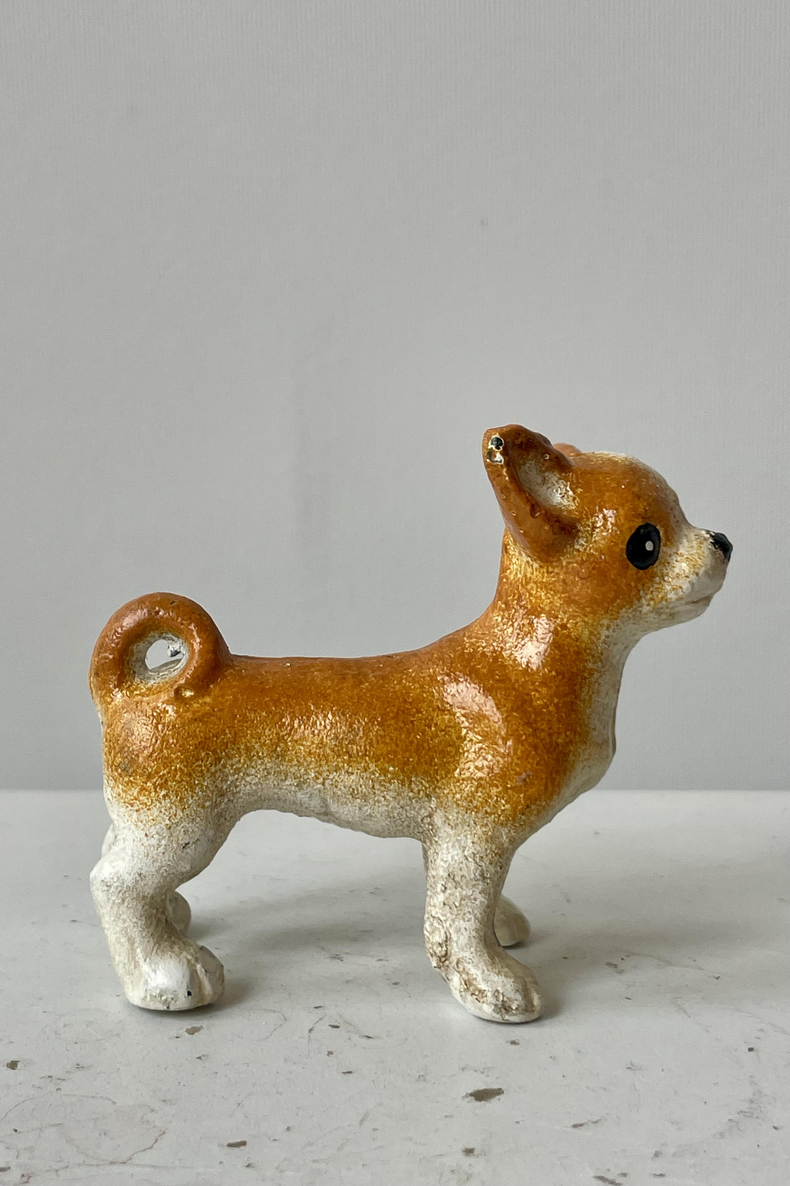 Photo of a white and tan Chihuahua figurine looking toward the right. The photo is taken from the side. It is shown on a white surface against a white wall.