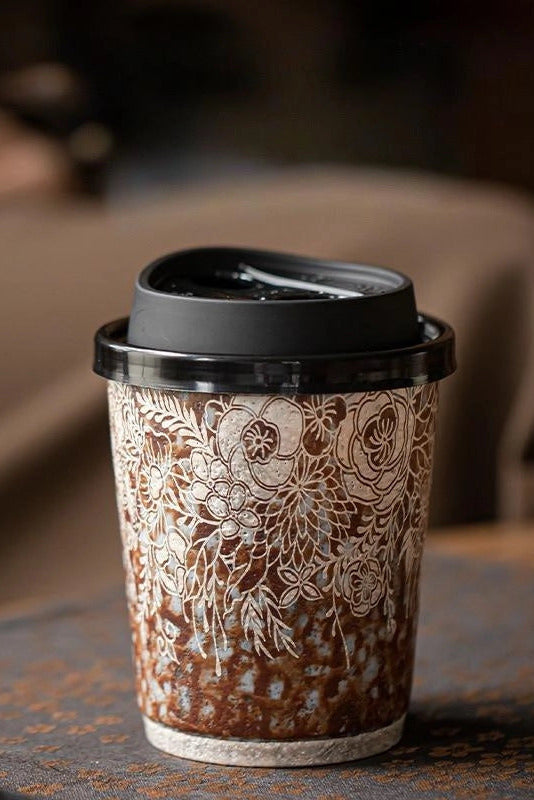 A reusable ceramic coffee cup with a black lid, decorated with an intricate white floral pattern over a brown base, sitting on a rustic table with a blurred background. ©Haku