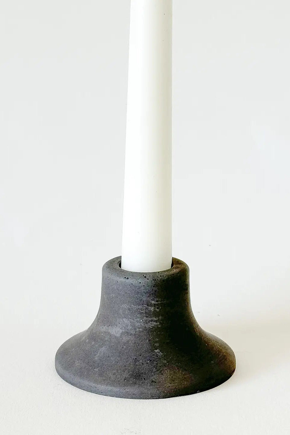 A dark charcoal gray ceramic candle holder with a flared base, holding a white taper candle, set against a plain white background. ©Sunday Nomad