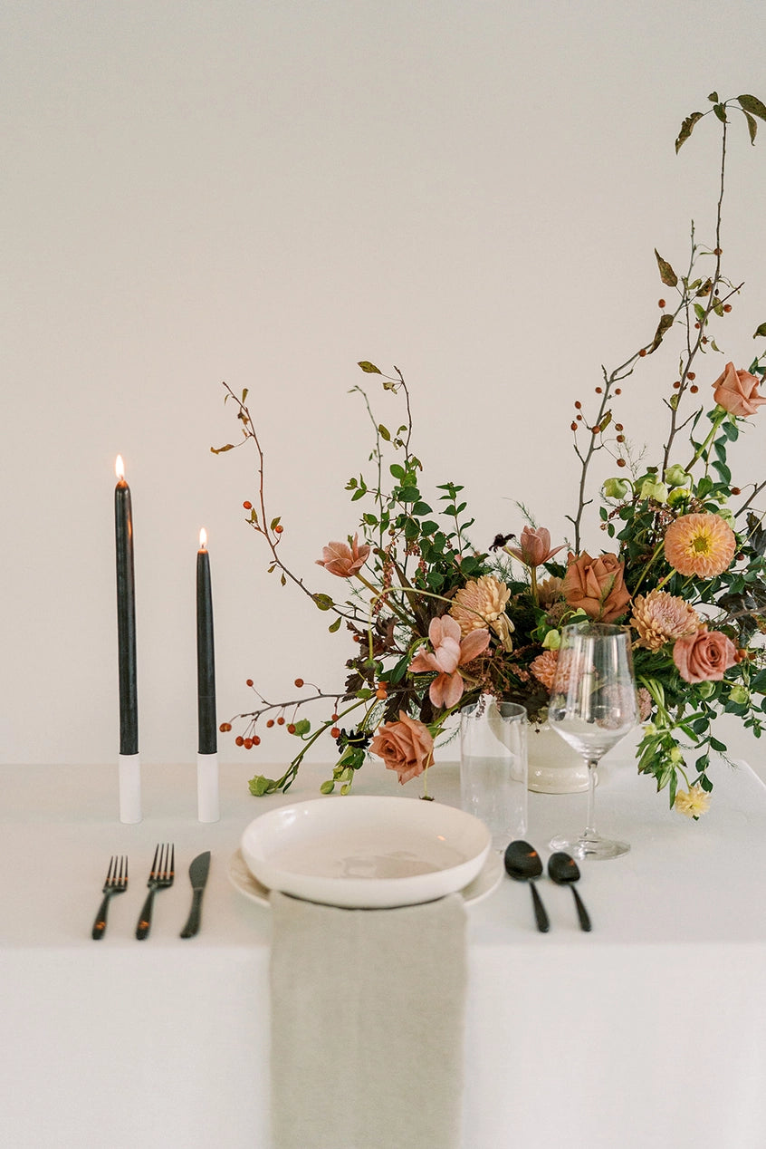 Mo & Co Charcoal beeswax taper candles shown lit and in a table setting with fruit and flowers.