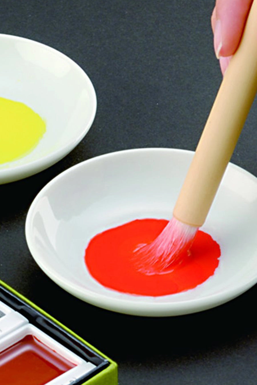 Person dipping a brush into red watercolor paint on a white plate with a black background ©Chartpak, Inc.