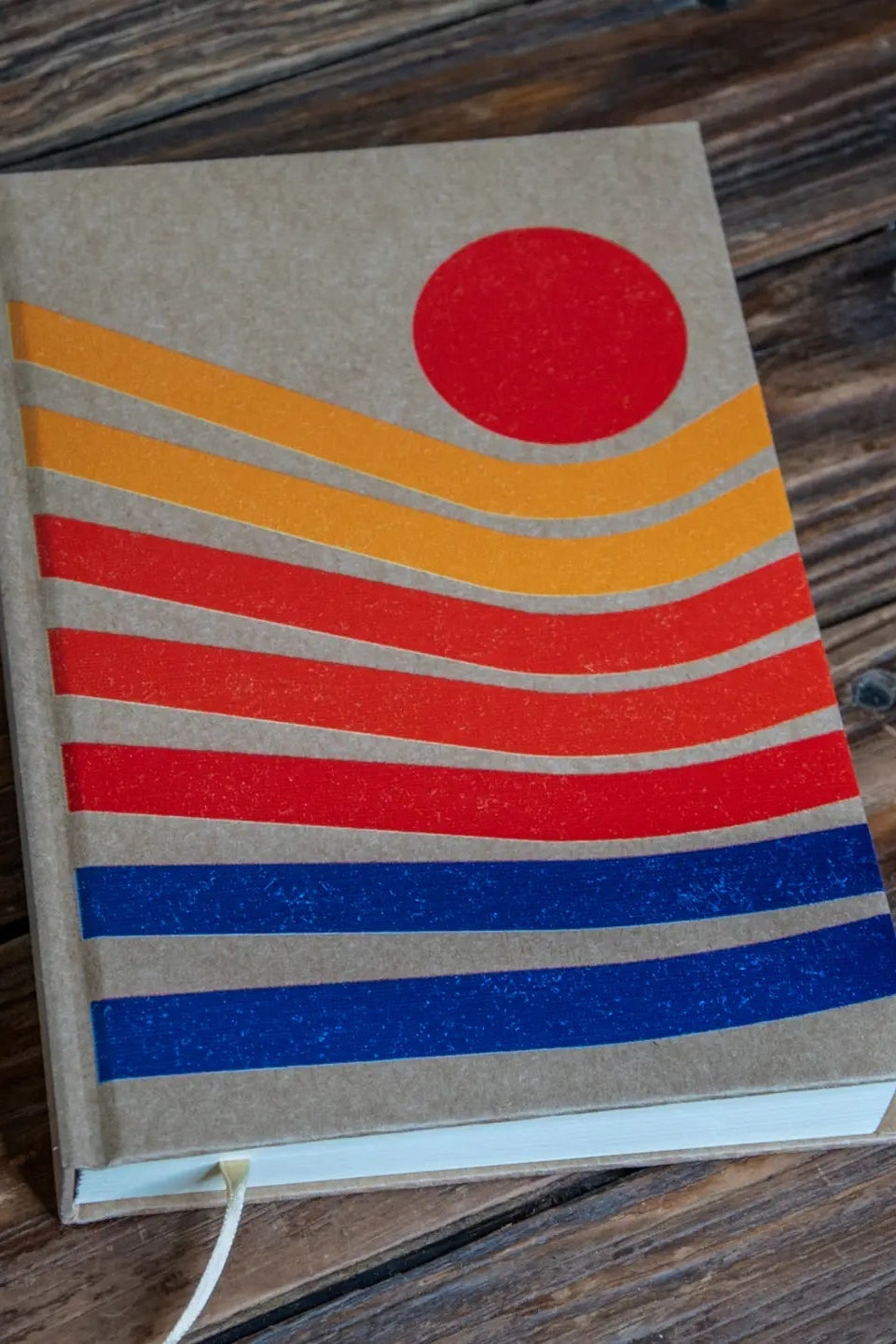 Sunset Notebook with a red, orange, and blue striped design on a wooden surface with a pen next to it. ©Glum Lot Press