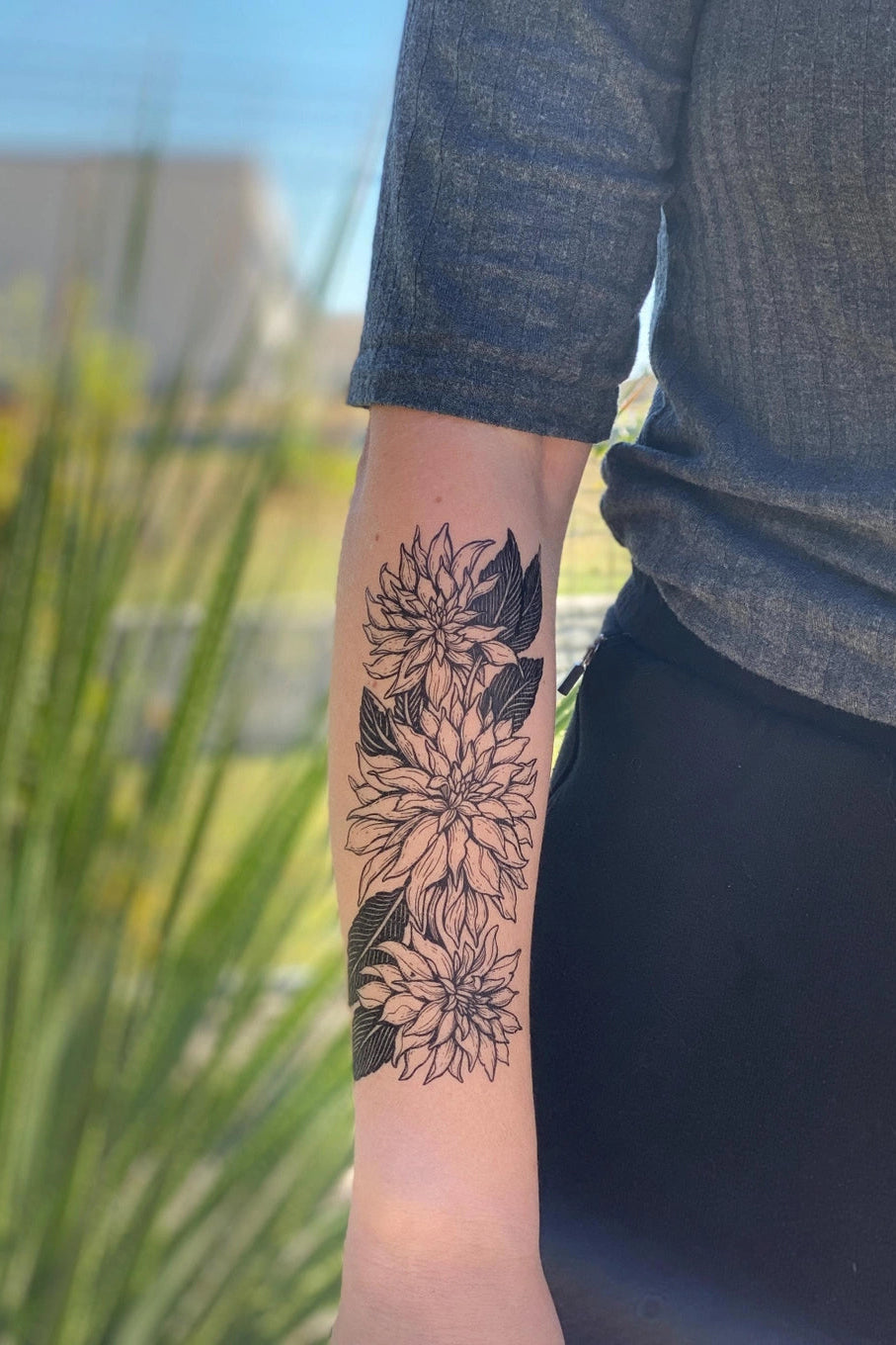 Person with a Peonies floral tattoo on their arm, standing outdoors with greenery in the background. ©Nature Tats