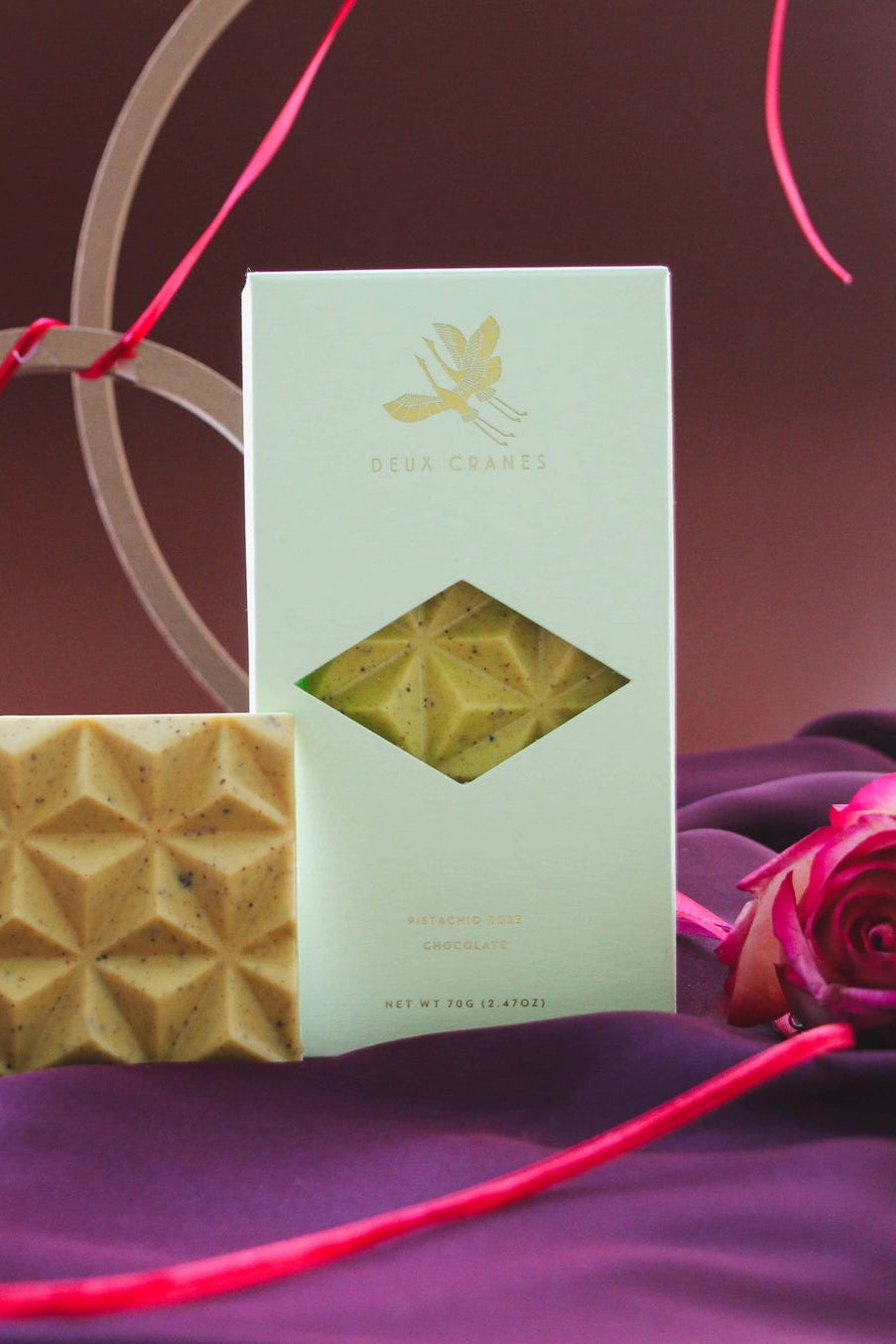 Geometric-patterned Pistachio Chocolate bar with packaging on a purple surface with pink roses. ©Deux Cranes
