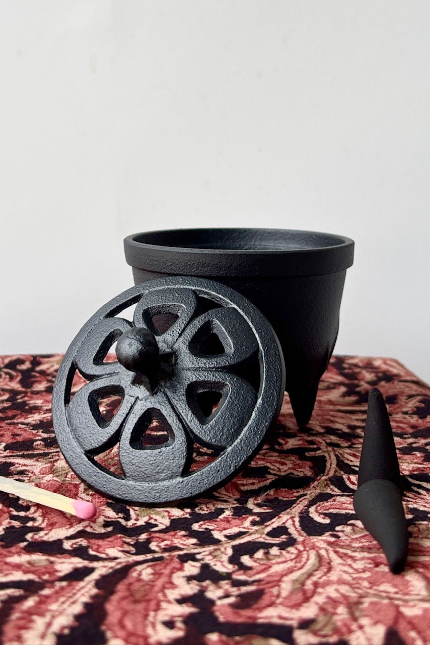 Cast Iron Incense charger and lid with a cut out floral pattern on a patterned tablecloth with incense cones and match ©Sprout Home
