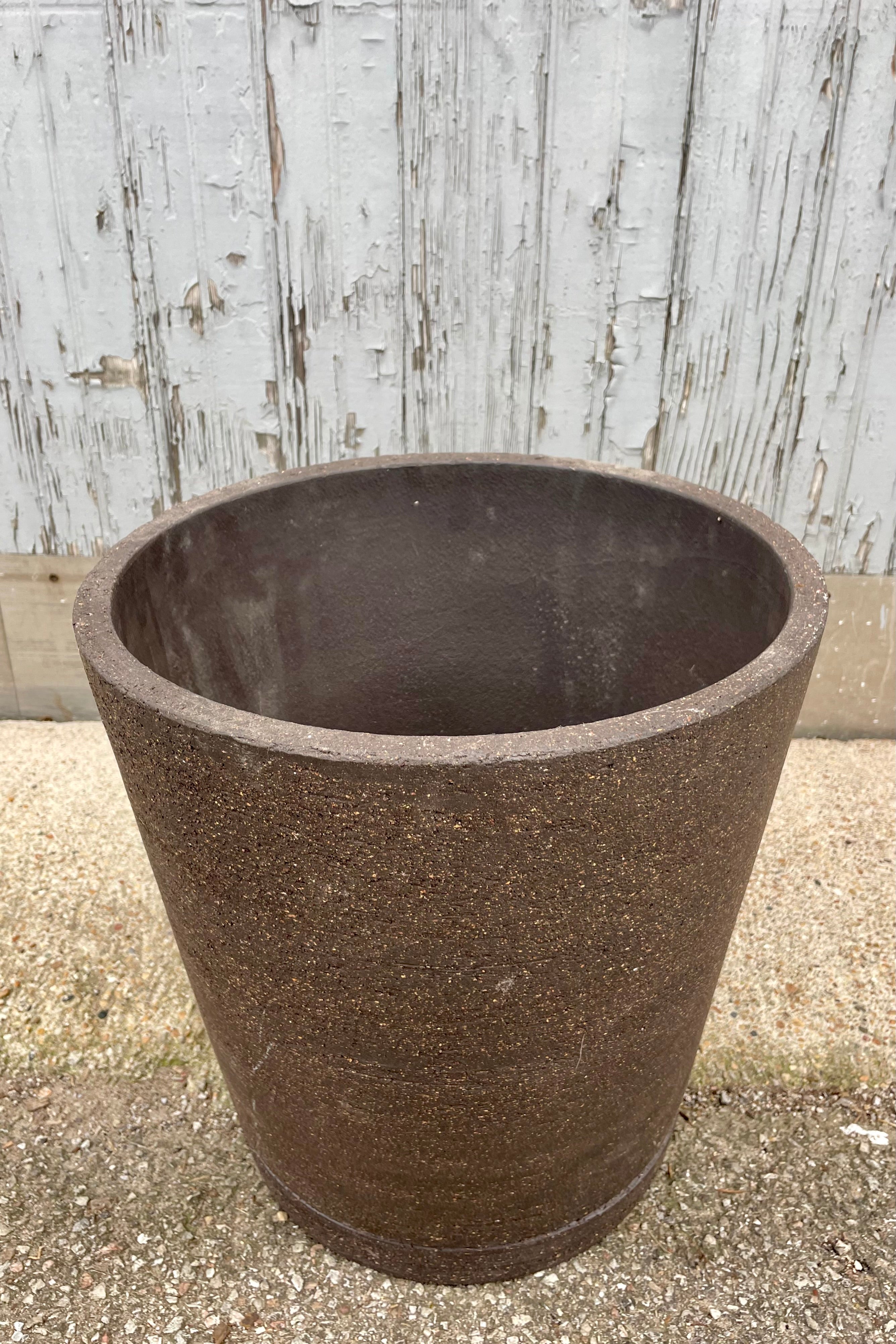 View from above. Peat colored I/O tapered planter in large showing the rough texture of the brown clay. Shown against a gray wall and cement floor.