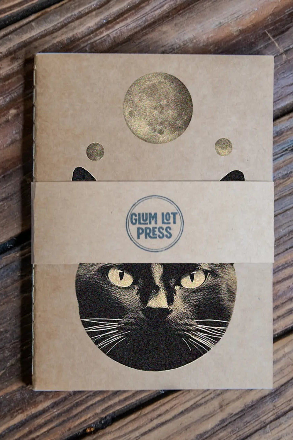 Notebook with cat design as part of the This is Surreal Pack Travel Journal 3 Pack Notebooks on wooden surface, featuring 'Glum Lot Press' branding. ©Glum Lot Press