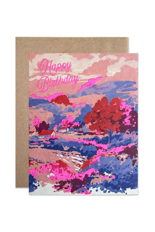 Birthday Countryside By Helen Dealtry Card with a scenic landscape illustration in pink, blue, red, and purple tones, with 'Happy Birthday' written in pink script at the top, shown with a brown kraft envelope behind it on a white background. ©Hartland Cards