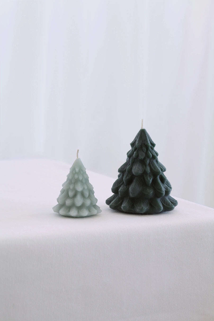 Two Christmas tree-shaped candles on a white surface with a light gray background ©Mo&Co.