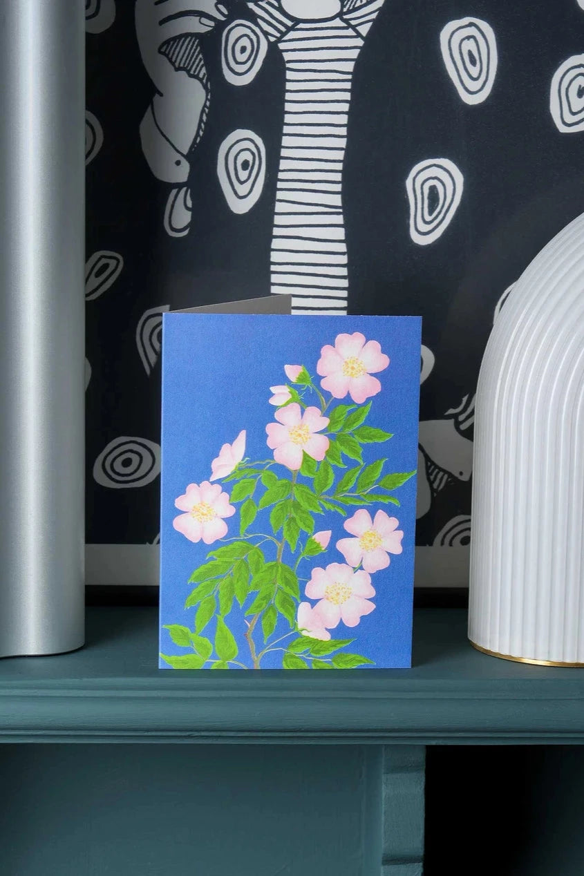 Dog rose greeting card by Stengun Drawings standing on a shelf with a black and white background.