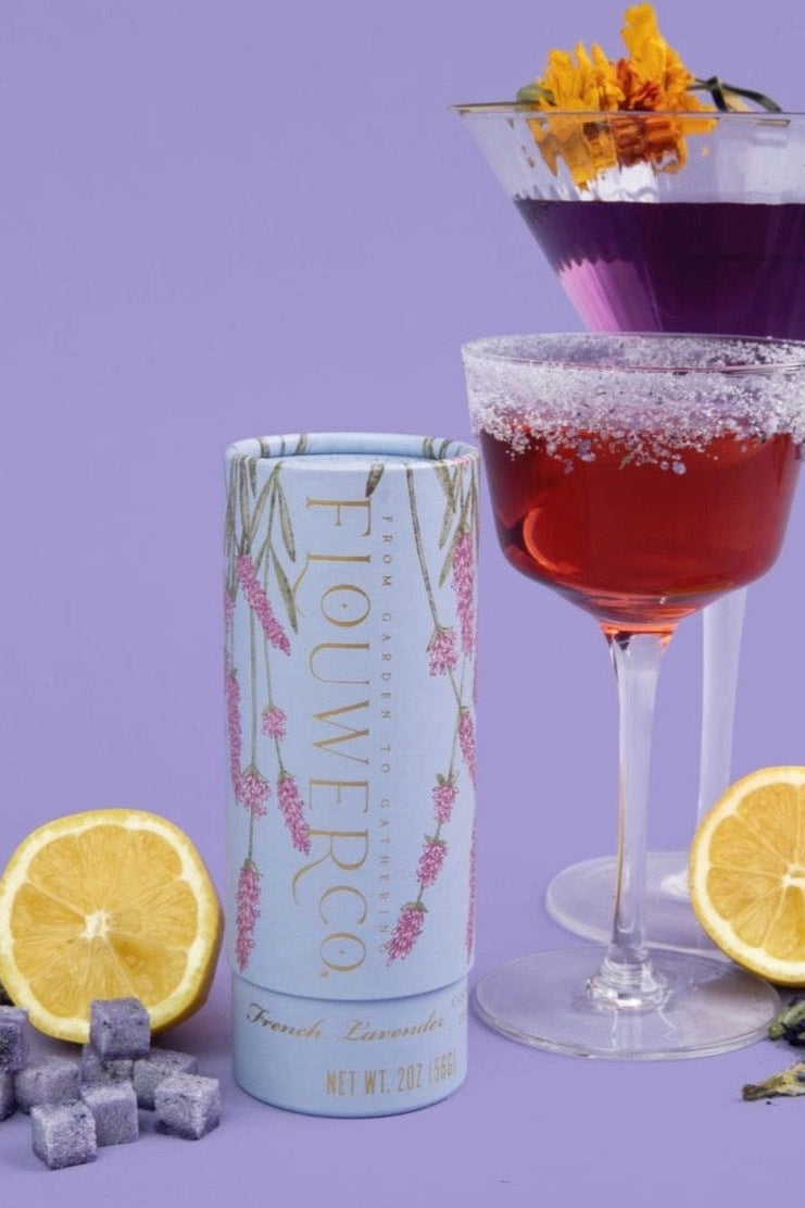 Two cocktails in glassware with a canister of lavender sugar cubes on a purple background ©Flouwer Co.