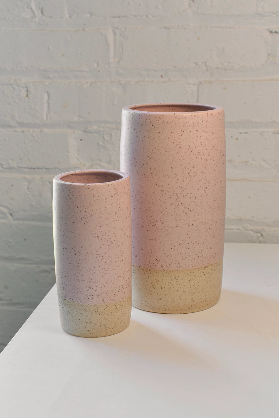 Two ceramic pink speckle Jacqueline vases with speckled finish on a white surface against a light gray brick wall. ©Little Fire Ceramics