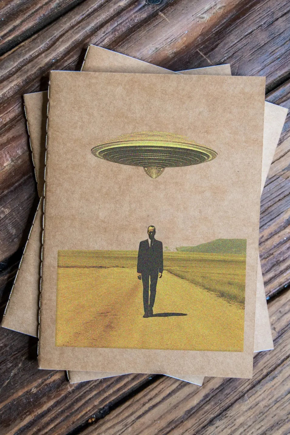 Notebook with a UFO and person illustration as part of the This is Surreal Pack Travel Journal 3 Pack Notebooks on a wooden surface ©Glum Lot Press