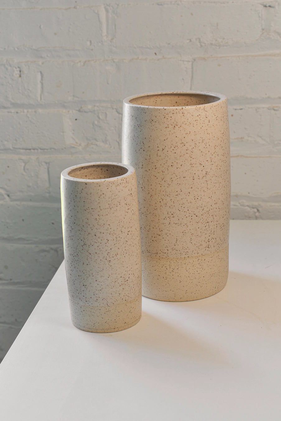 Two ceramic cream speckle Jacqueline vases on a white surface with a light gray brick wall background ©Little Fire Ceramics