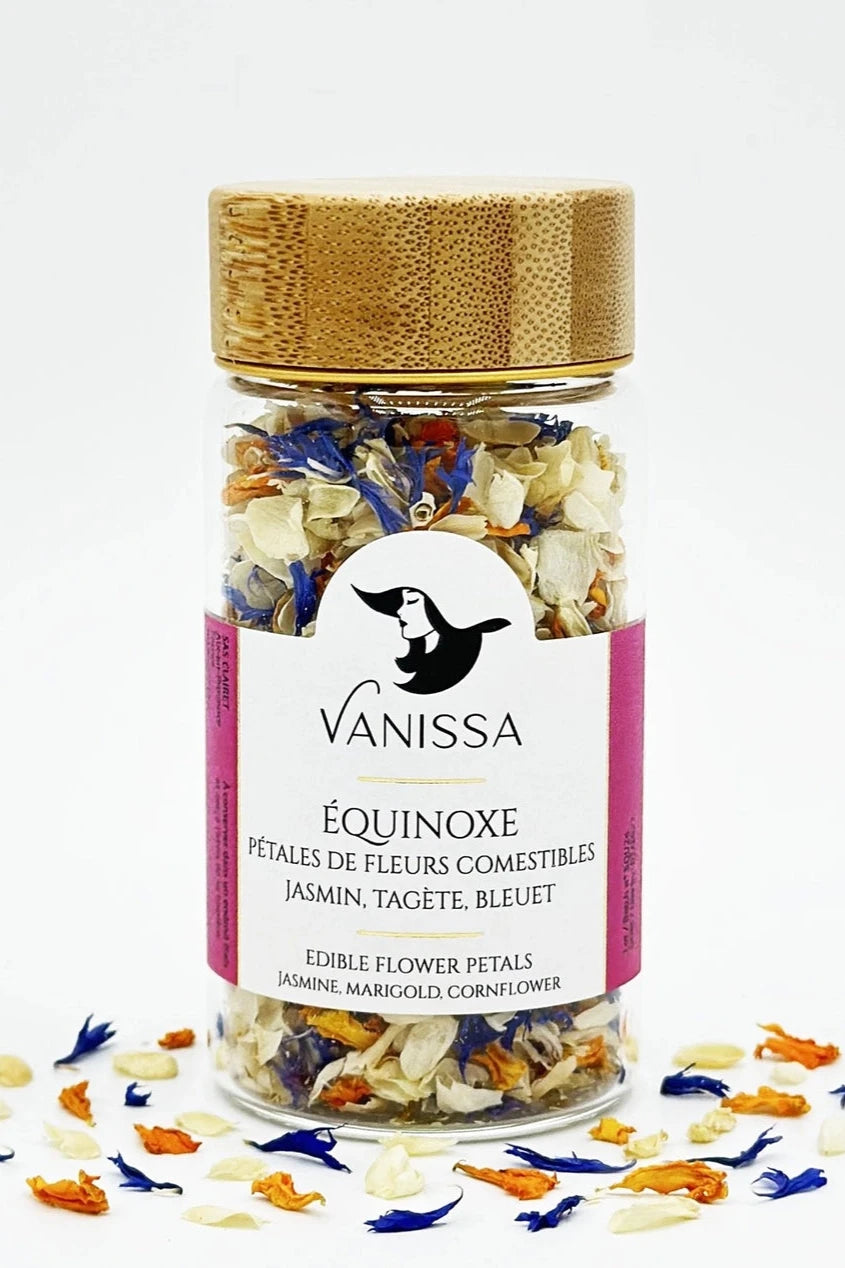Equinoxe edible flower petals by Vanissa showing its glass and wood tube packaging with loose leaves around it.