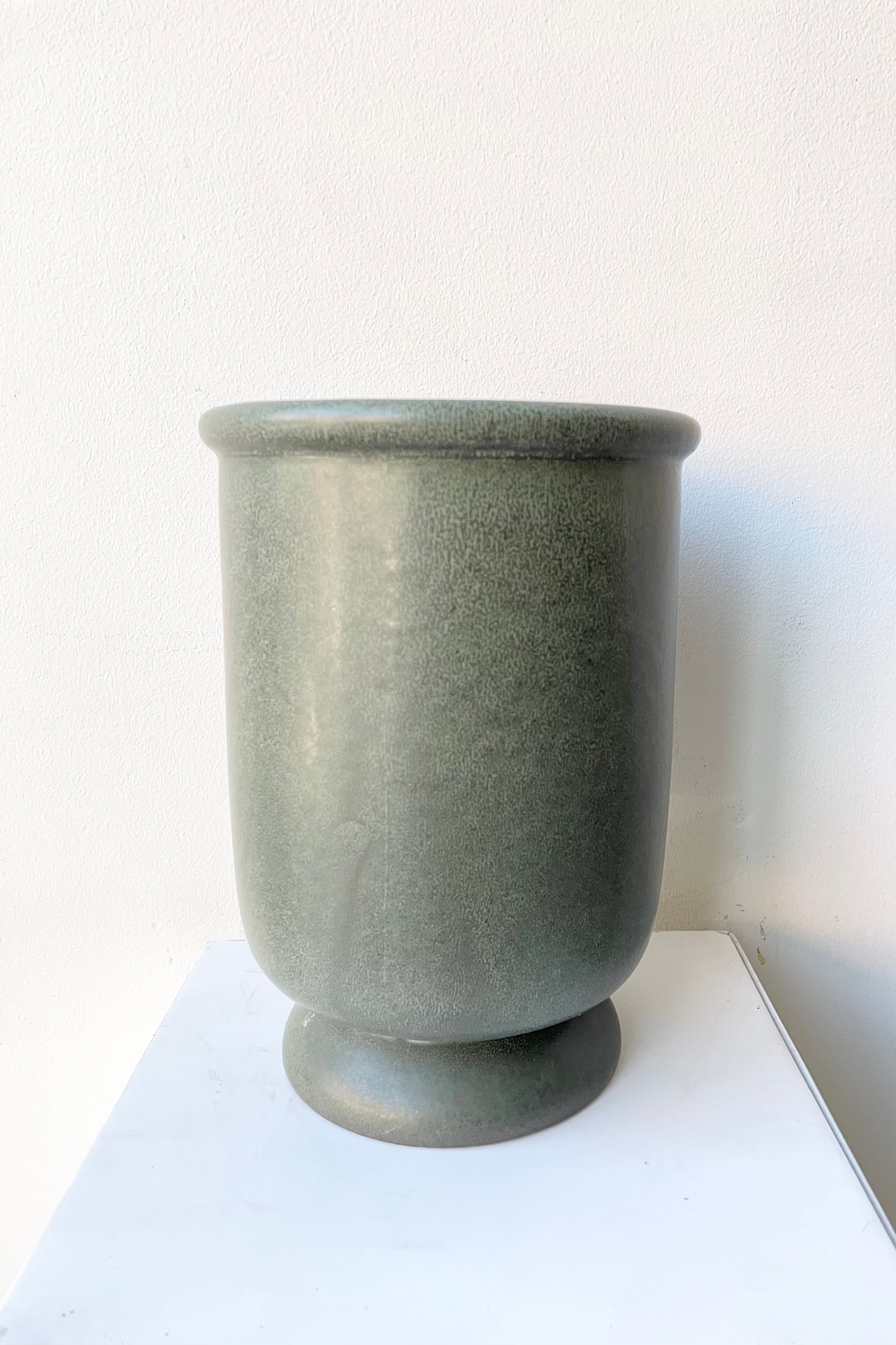 The dark green stoneware urn vase shown from the side against a white wall.