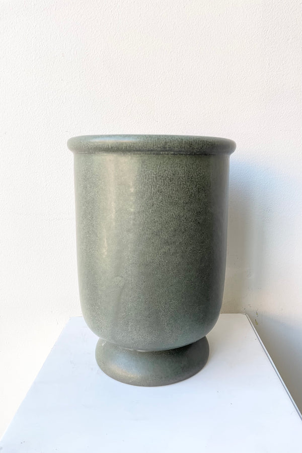 The dark green stoneware urn vase shown from the side against a white wall. 