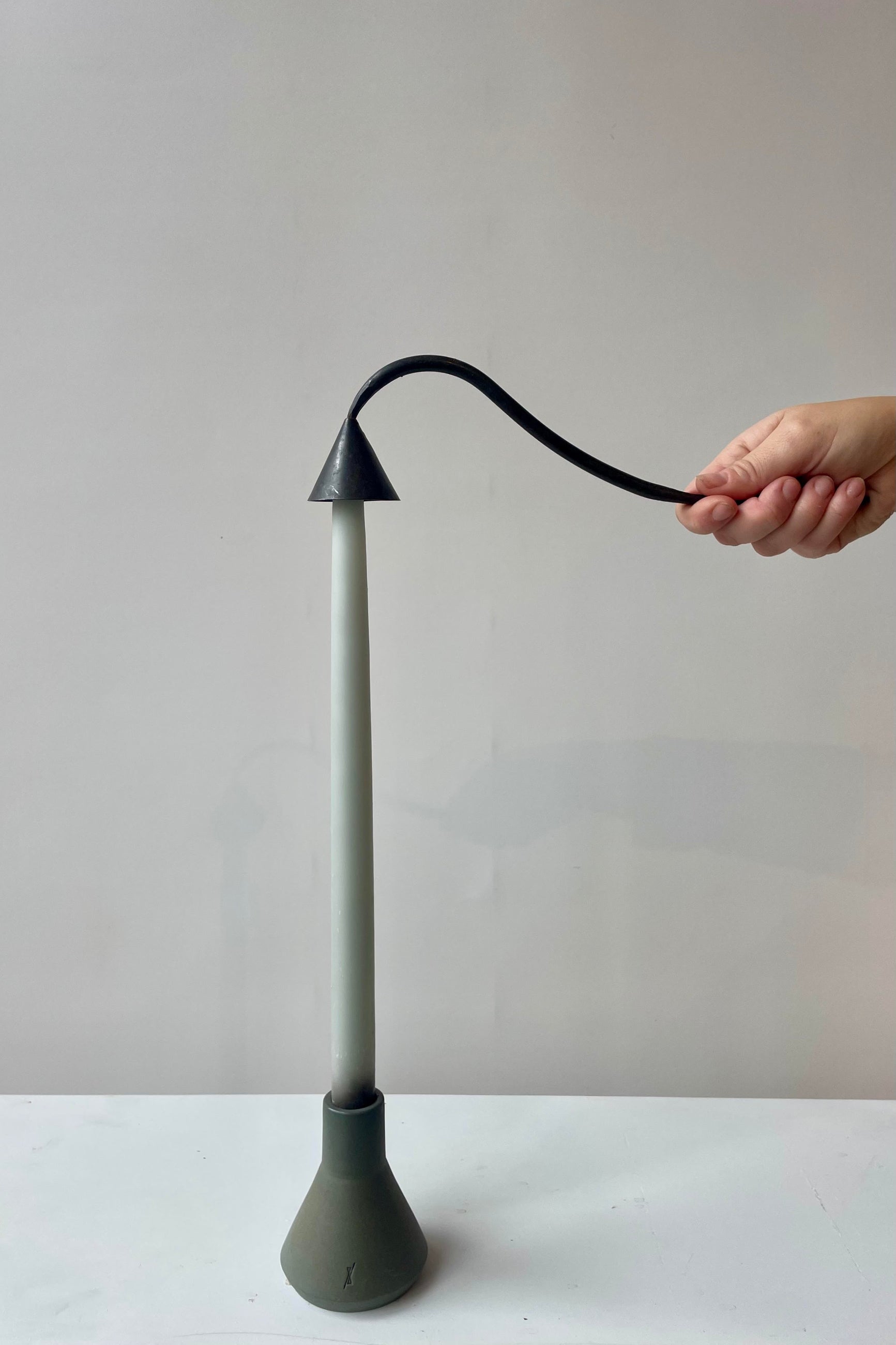 Photo of a hand holding a black metal candle snuffer in a white room. The snuffer is placed over the tip of a pale green taper candle that sits within a dark green candle holder on a white surface. ©Sprout Home