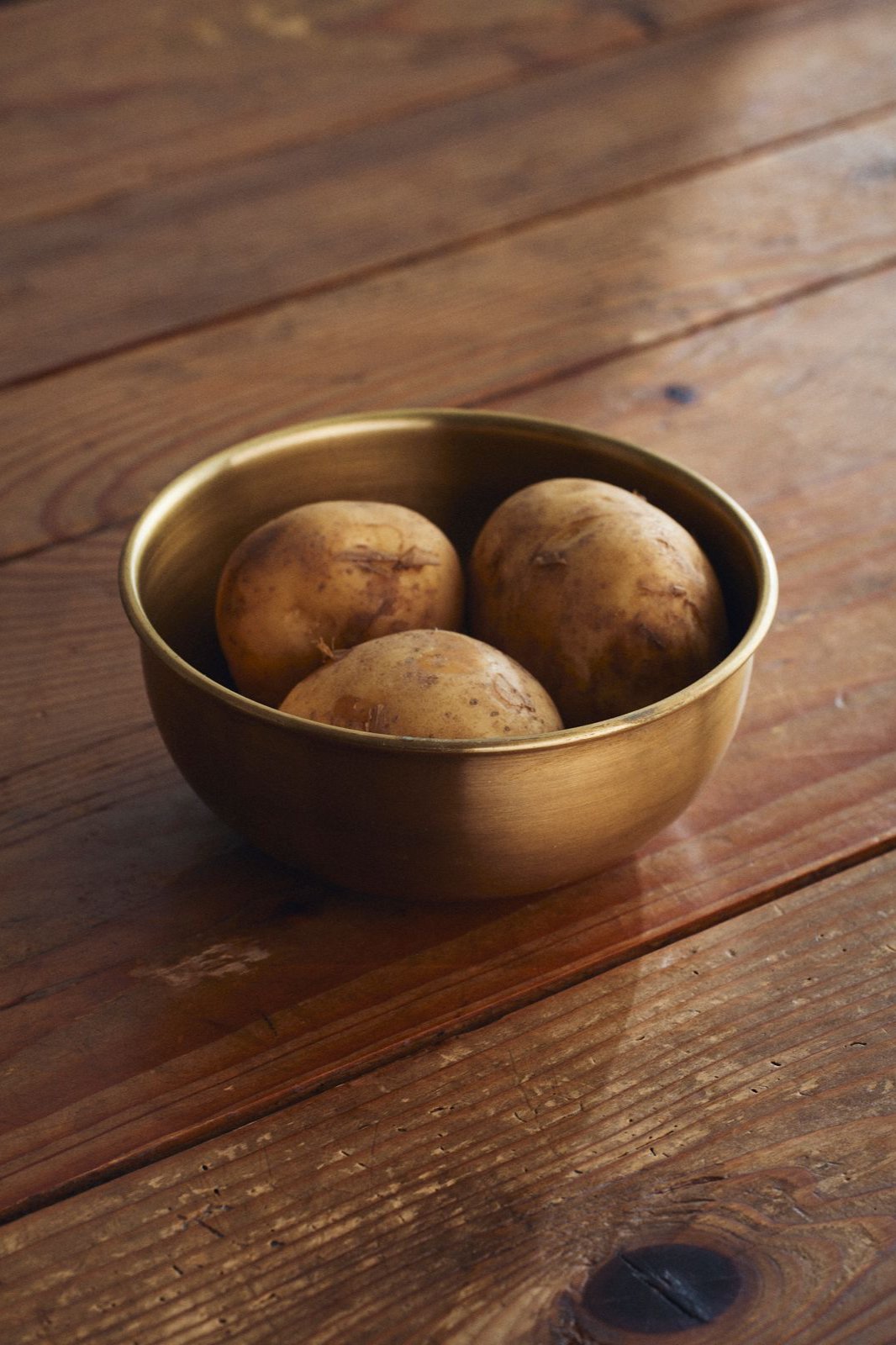 Large Brass Bowl by Fog Linen Work with potatoes inside and sitting on a wood table.