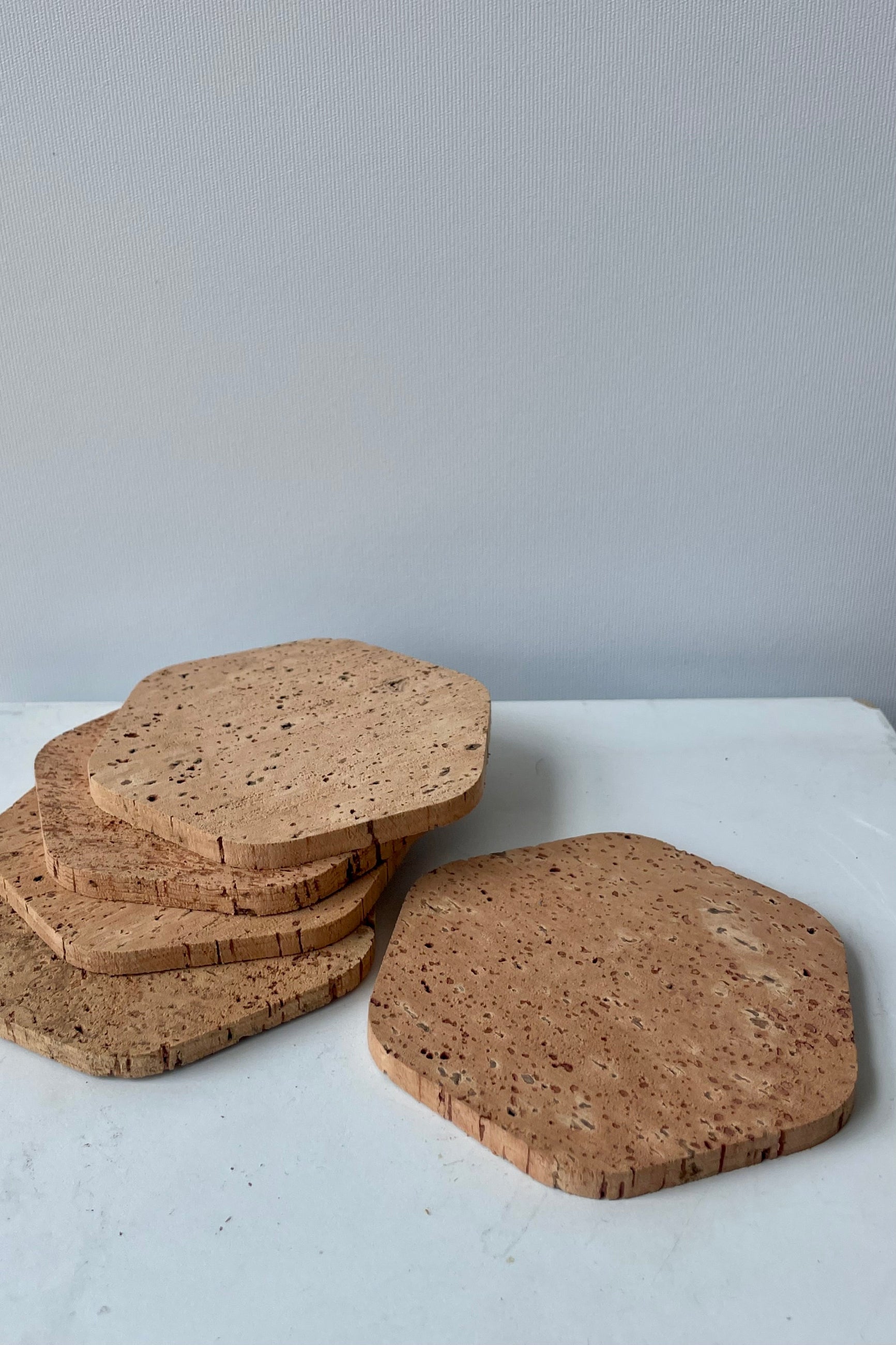 A set of five six-sided cork coasters are arranged in a spiraled stack of four with one individual coaster ahead of the rest. These polygon cork coasters are photographed on a white table against a white wall. ©Sprout Home
