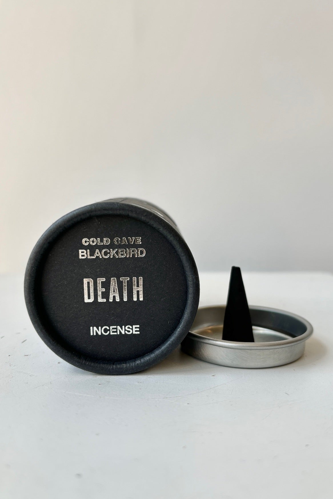 A black cardboard cylinder featuring silver patterning and type with a circular silver tray and one cone incense on a white table with white background. This scent is called Death and the fragrance contains notes of leather, marble and sage. ©Sprout Home