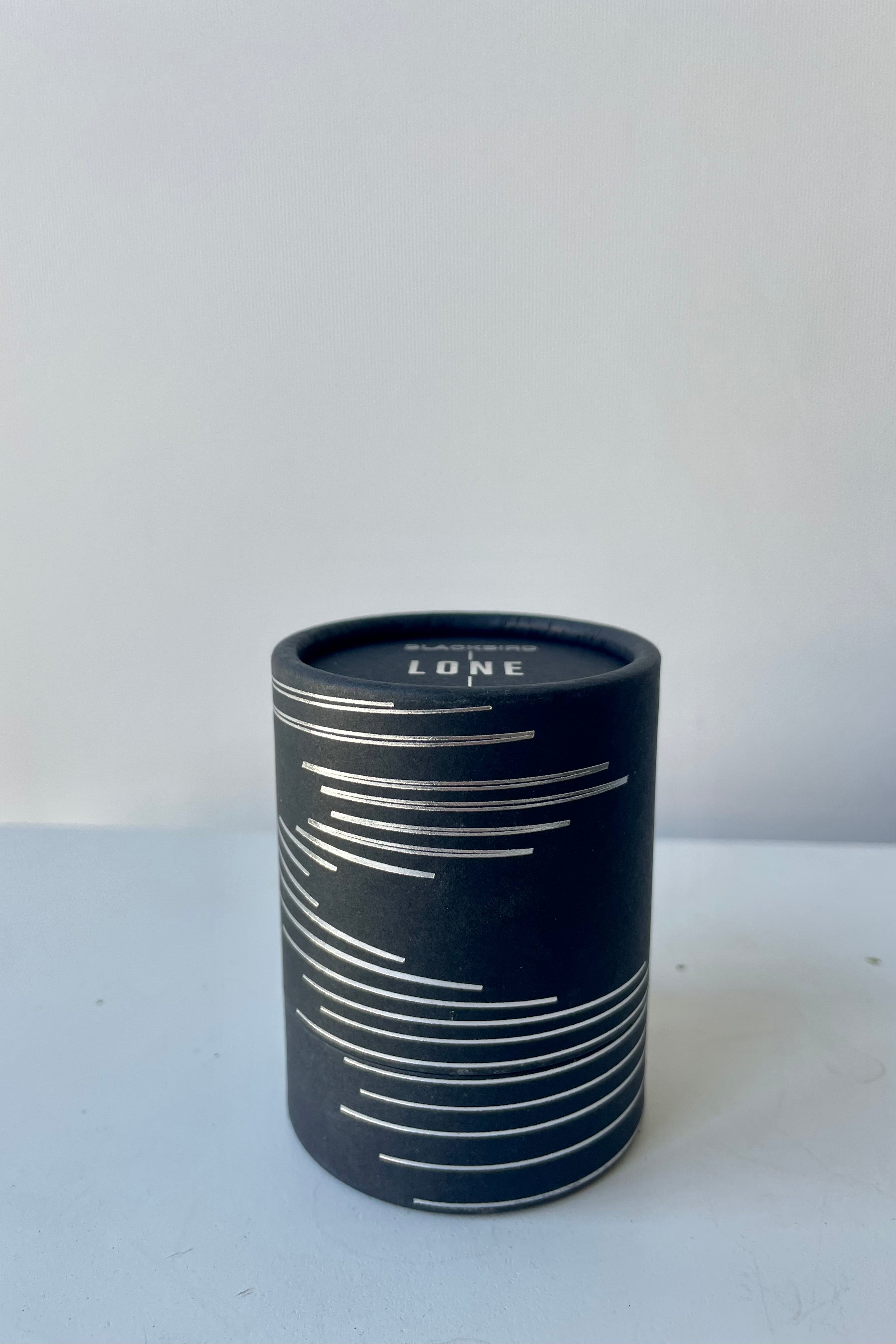 A black cylinder of Blackbird incense sits on a white surface in a white room. This incense is called Lone and the package features a silver lined design.