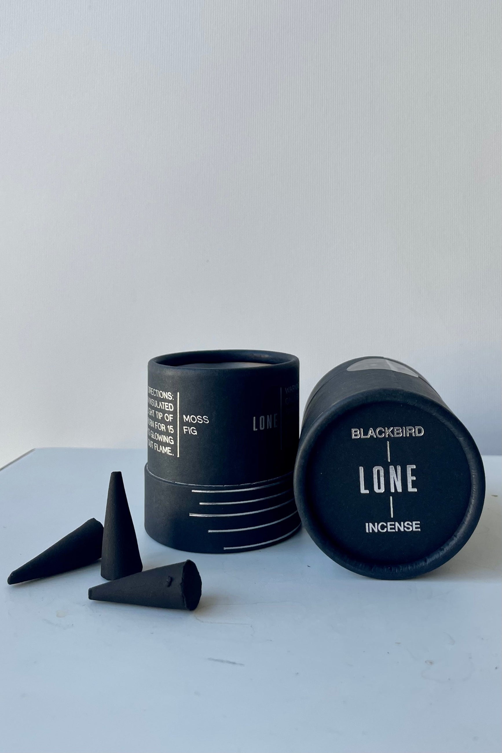 A black cylinder of Blackbird incense sits on a white surface in a white room. This incense is called Lone and the package features a silver lined design. To the left sits a scattering of black incense cones. ©Sprout Home