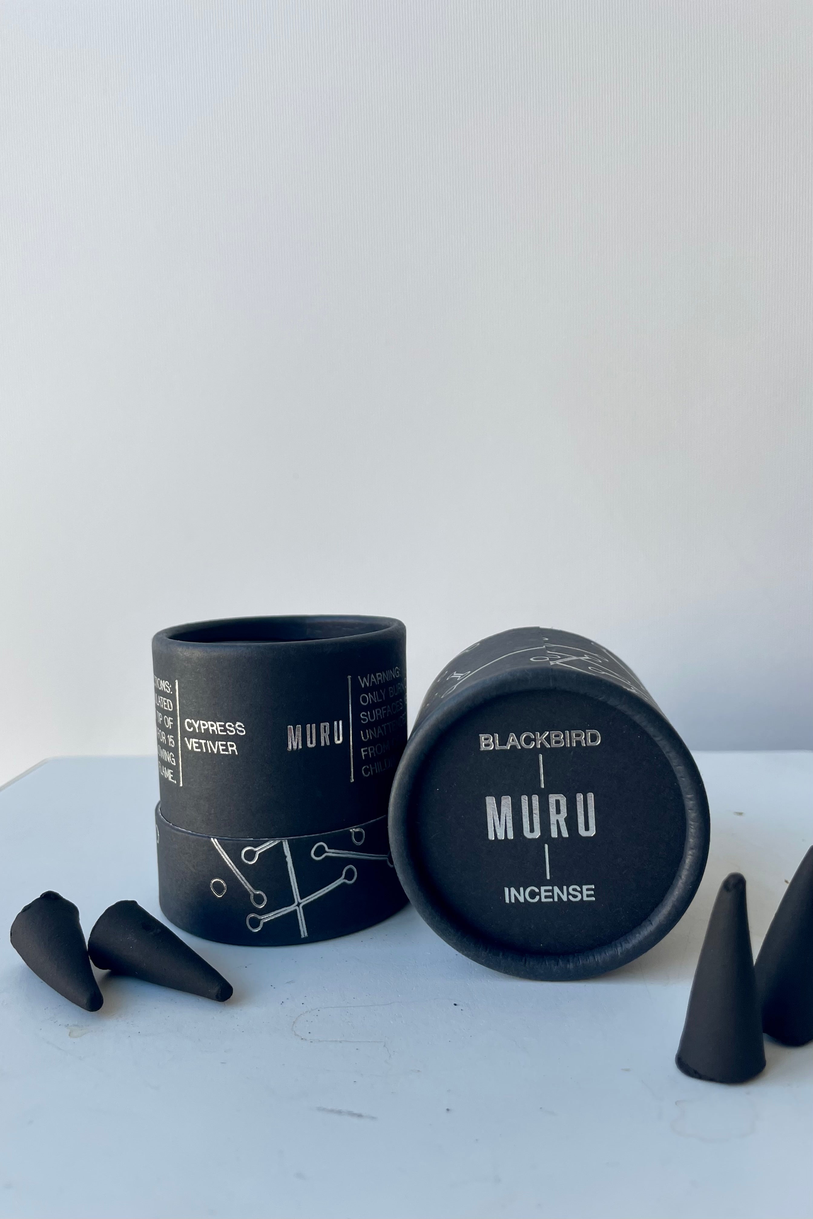 A black cylinder of Blackbird incense sits on a white table in a white room. The incense is called Muru. The packaging features a black background with silver lines and circles. To the left and right and black incense cones.
