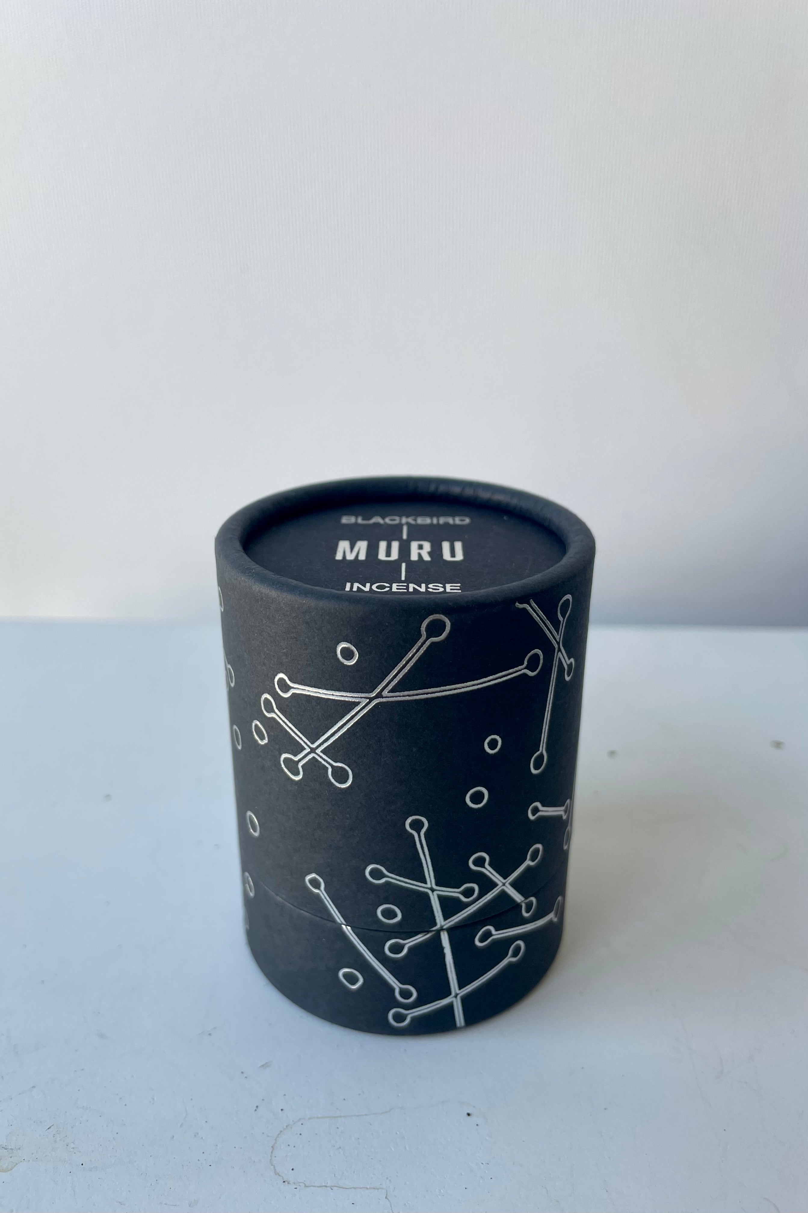 A black cylinder of Blackbird incense sits on a white table in a white room. The incense is called Muru. The packaging features a black background with silver lines and circles.
