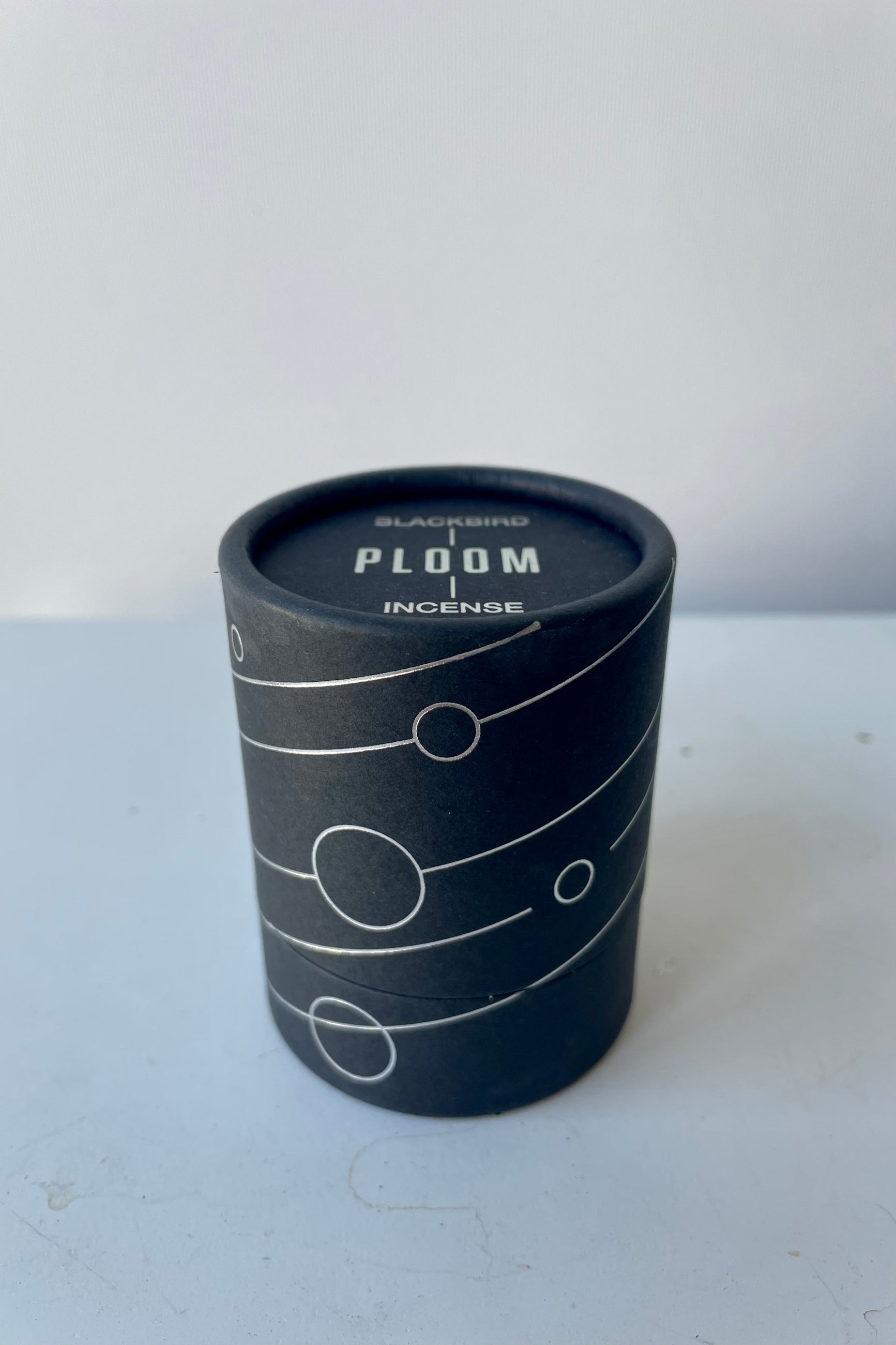 A black cylinder containing Blackbird Incense sits on a white table in a white room. The incense is called Ploom. Packaging features a black background with intersecting silver lines and circles. ©Sprout Home