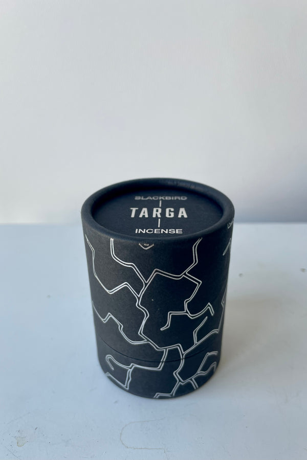 A black cylinder of Blackbird incense sits on a white table in a white room. The incense is called Targa. The packaging features parallel jagged silver lines.