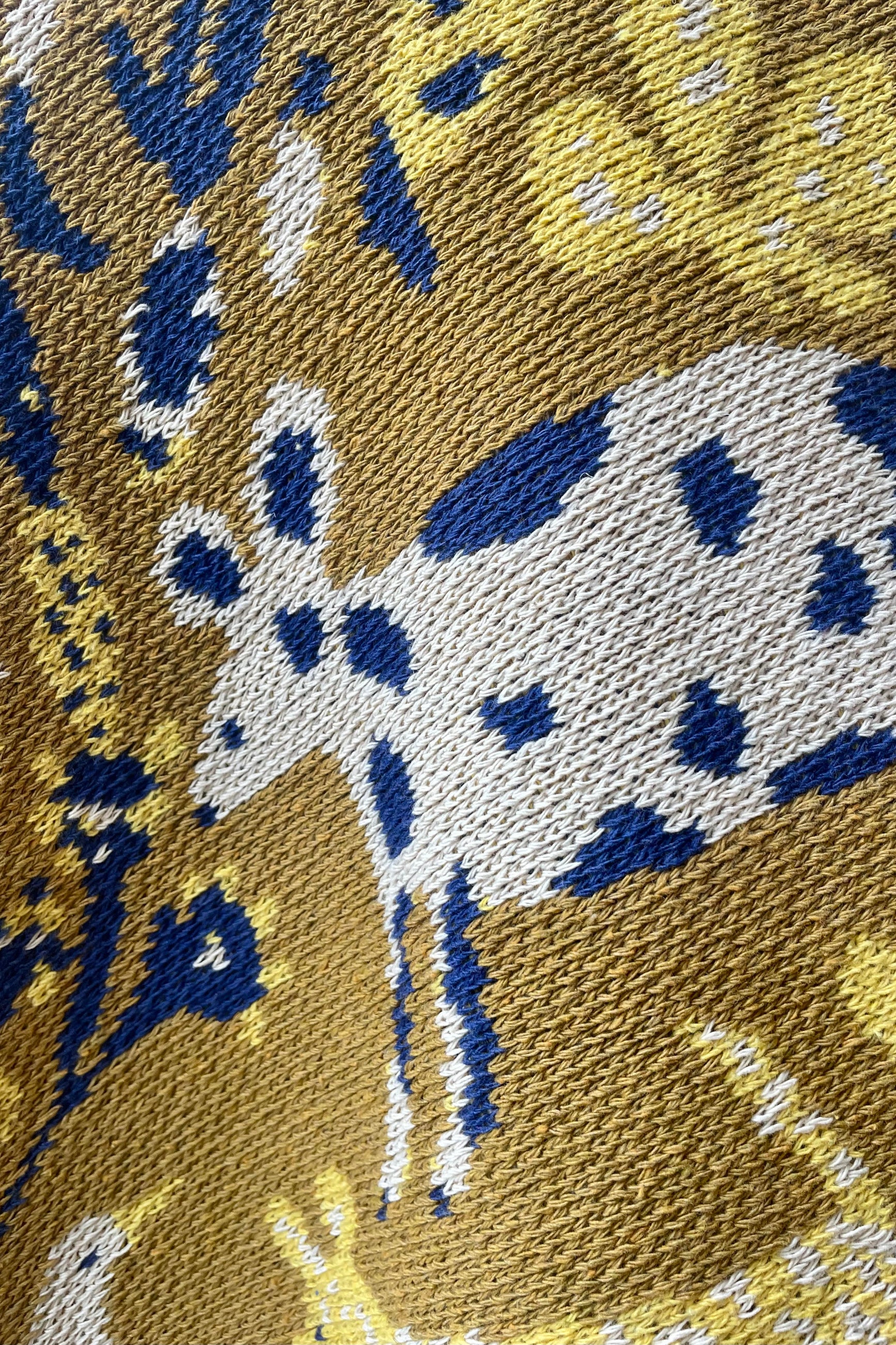 Close up photo of weave and character of Swells Ochre woven blankiet with golden and blue patterns against a white wall. ©Sprout Home