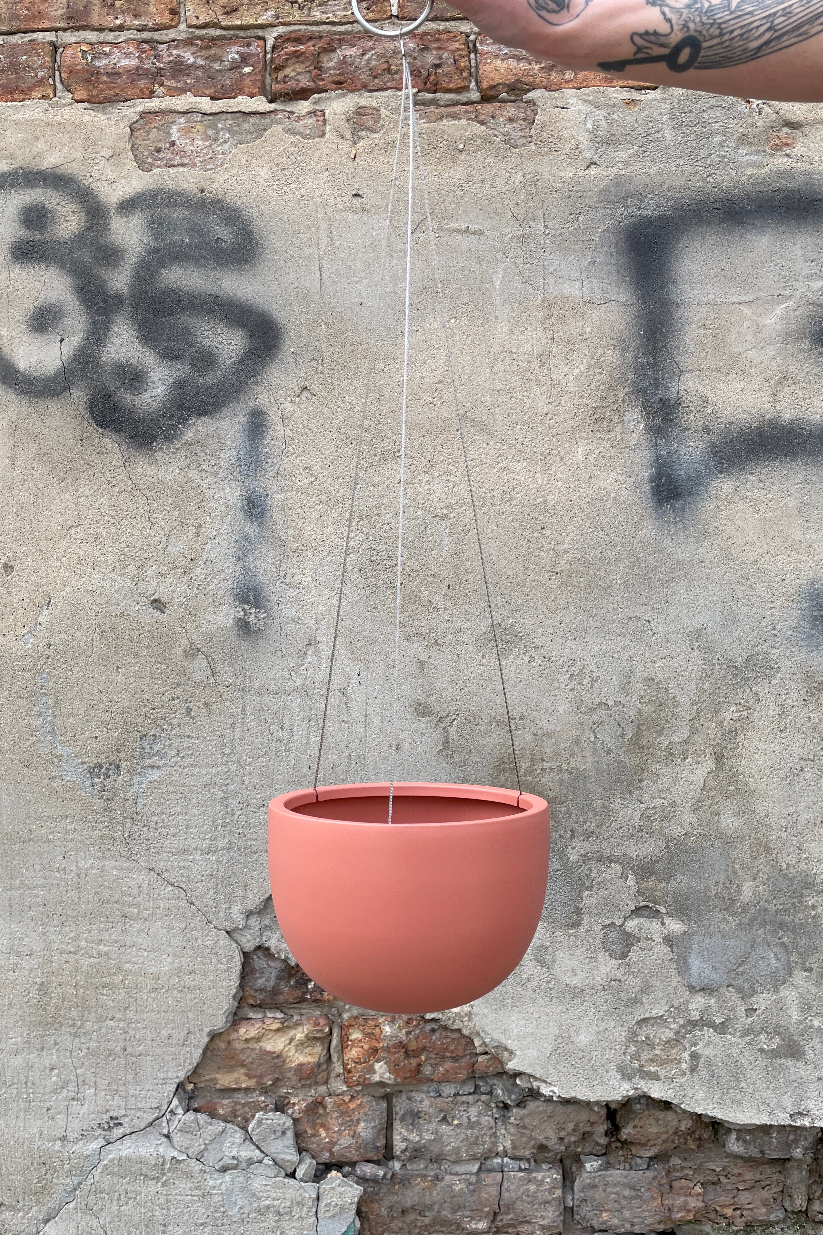 Clifton Matte Dusty Pink hanging pot small being held emtpy against a concrete and brick wall.
