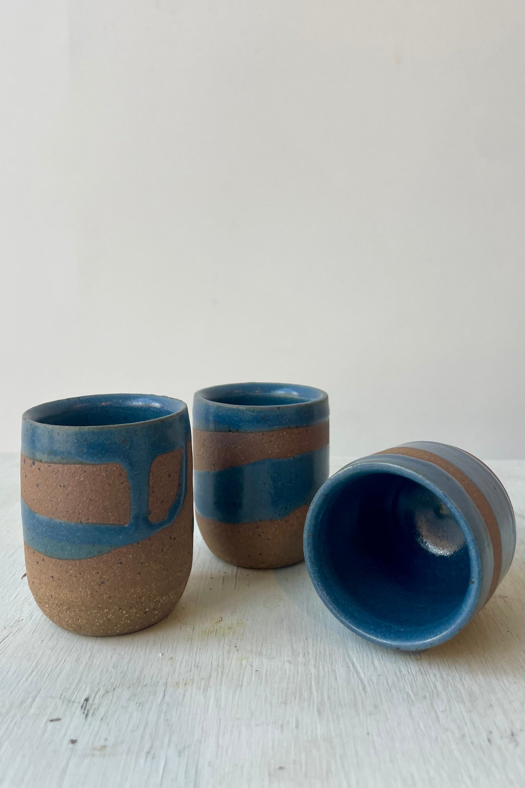 A trio of small ceramic cups with brown clay and a dark blue glaze in front of a white wall ©Sprout Home