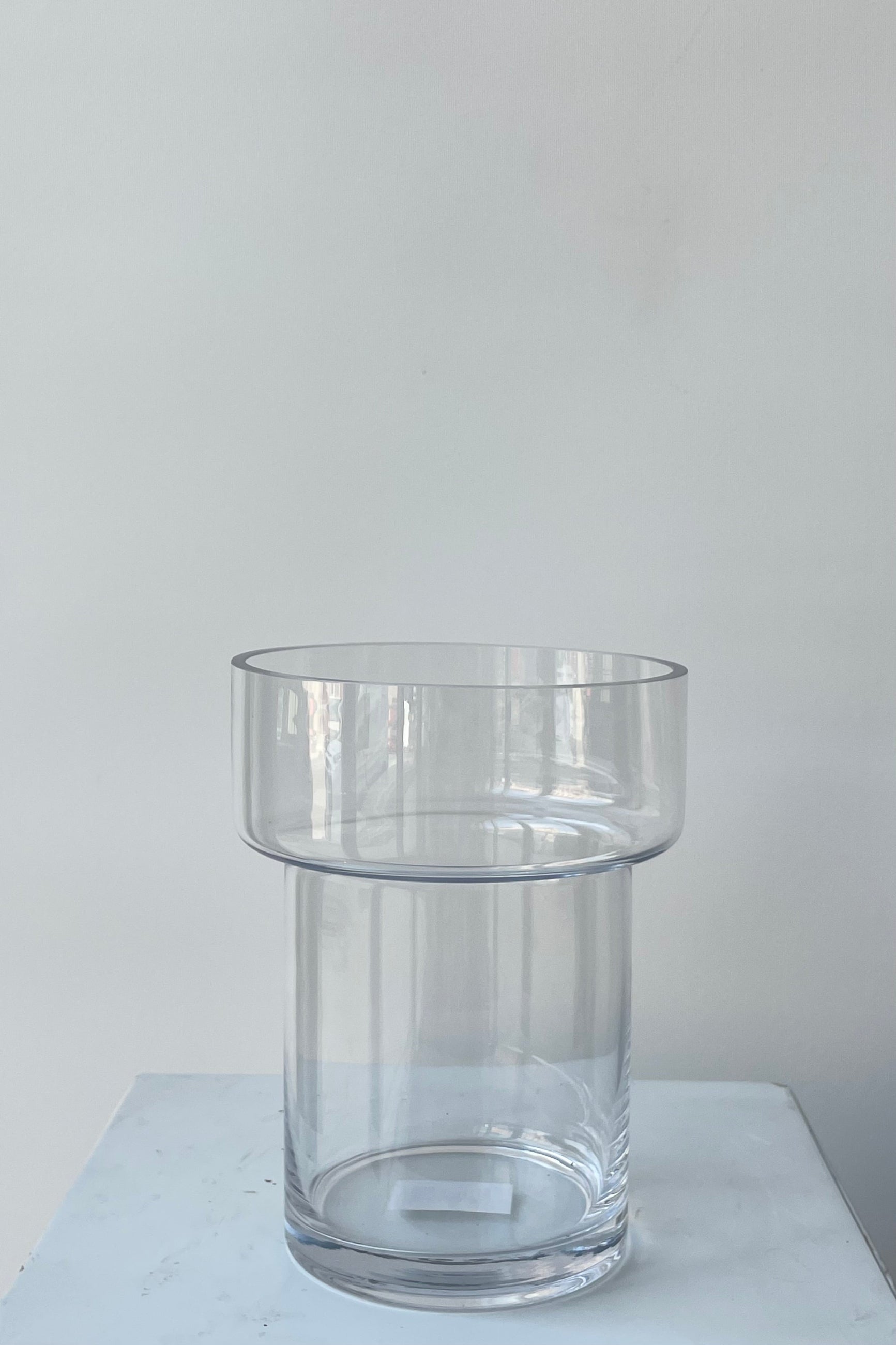 Photo of a clear glass Keeper vase. The vase is cylindrical with a wider top portion and a narrower two-thirds of its height. The vase is photographed in a white room against a white wall. ©Sprout Home