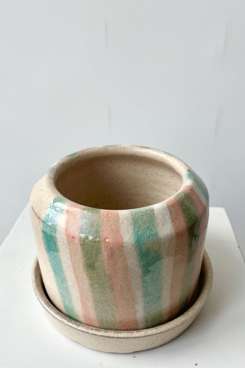 Photo of a glazed terra cotta flower pot and saucer. The planter is set within a saucer and both sit on a white surface in a white room. The semi-gloss glaze features irregular semi-opaque stripes in the glaze. The strapes are shades of coral, light blue and green. The phot is from above looking into the opening of the planter.