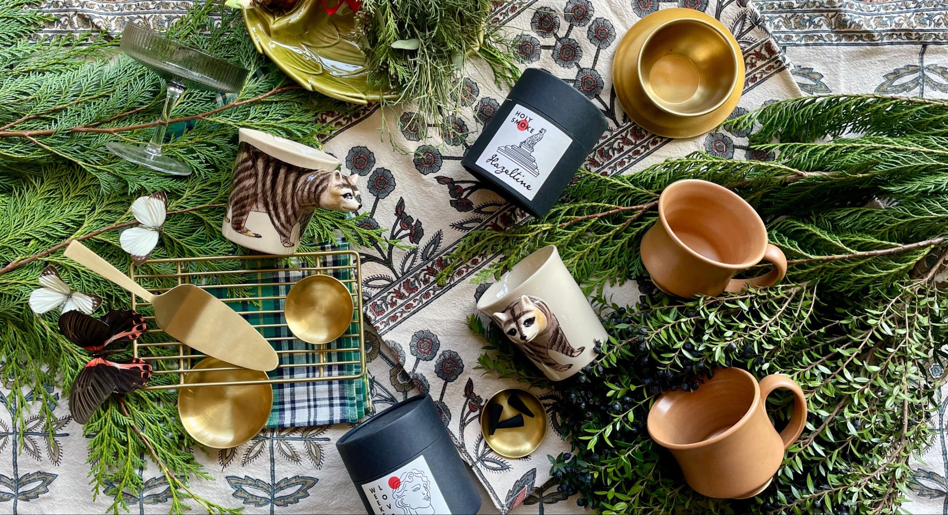 An overhead photo of gift items of brass and ceramic and candles displayed on a bed of evergreen foliage and mixed textiles. ©Sprout Home