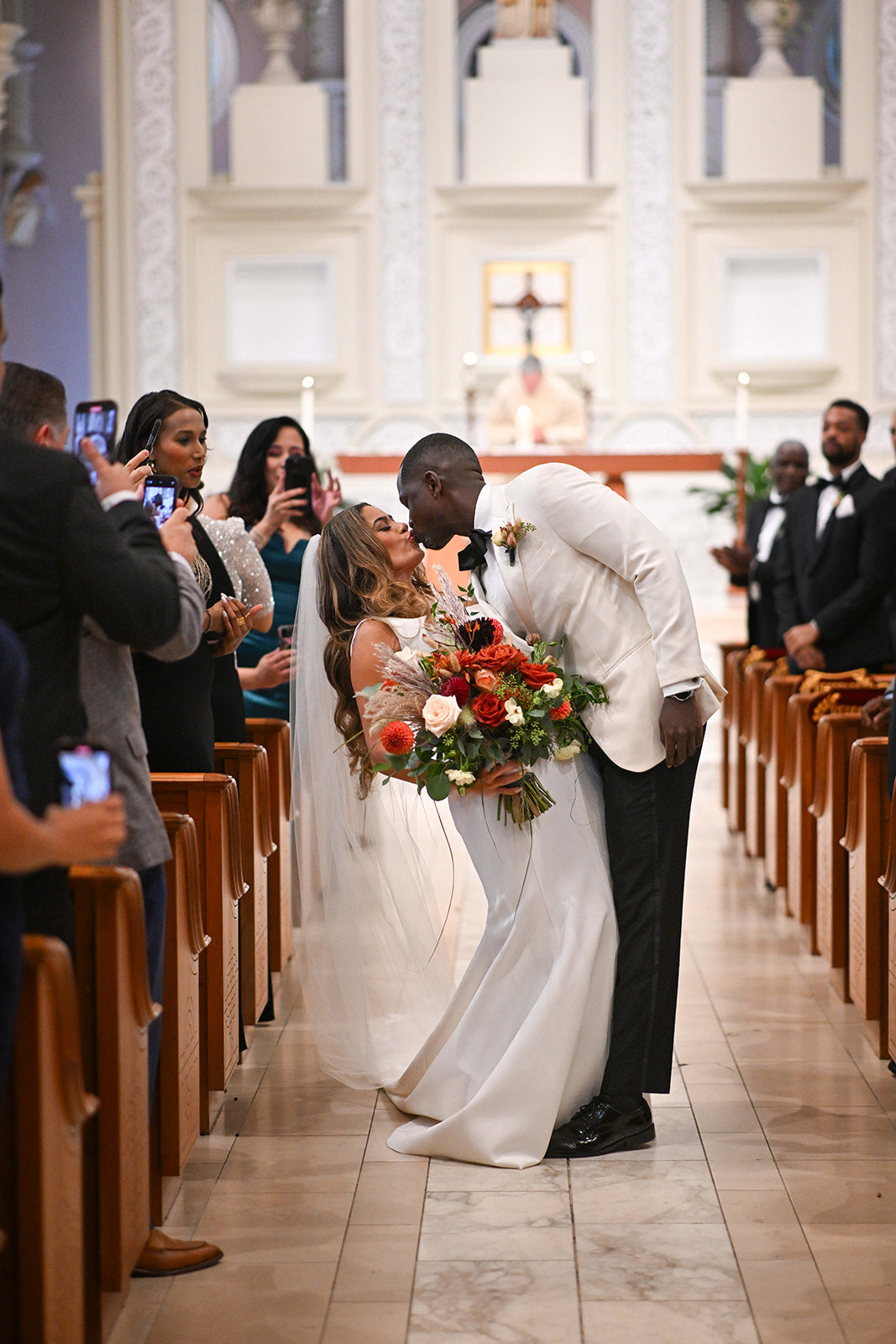 Sam & Kwaku's wedding. Bride holding a large earthy colored bouquet with huge flower pops standing and kissing in the aisle of a church. Floral by Sprout Home. Photograph by Mack Julion
