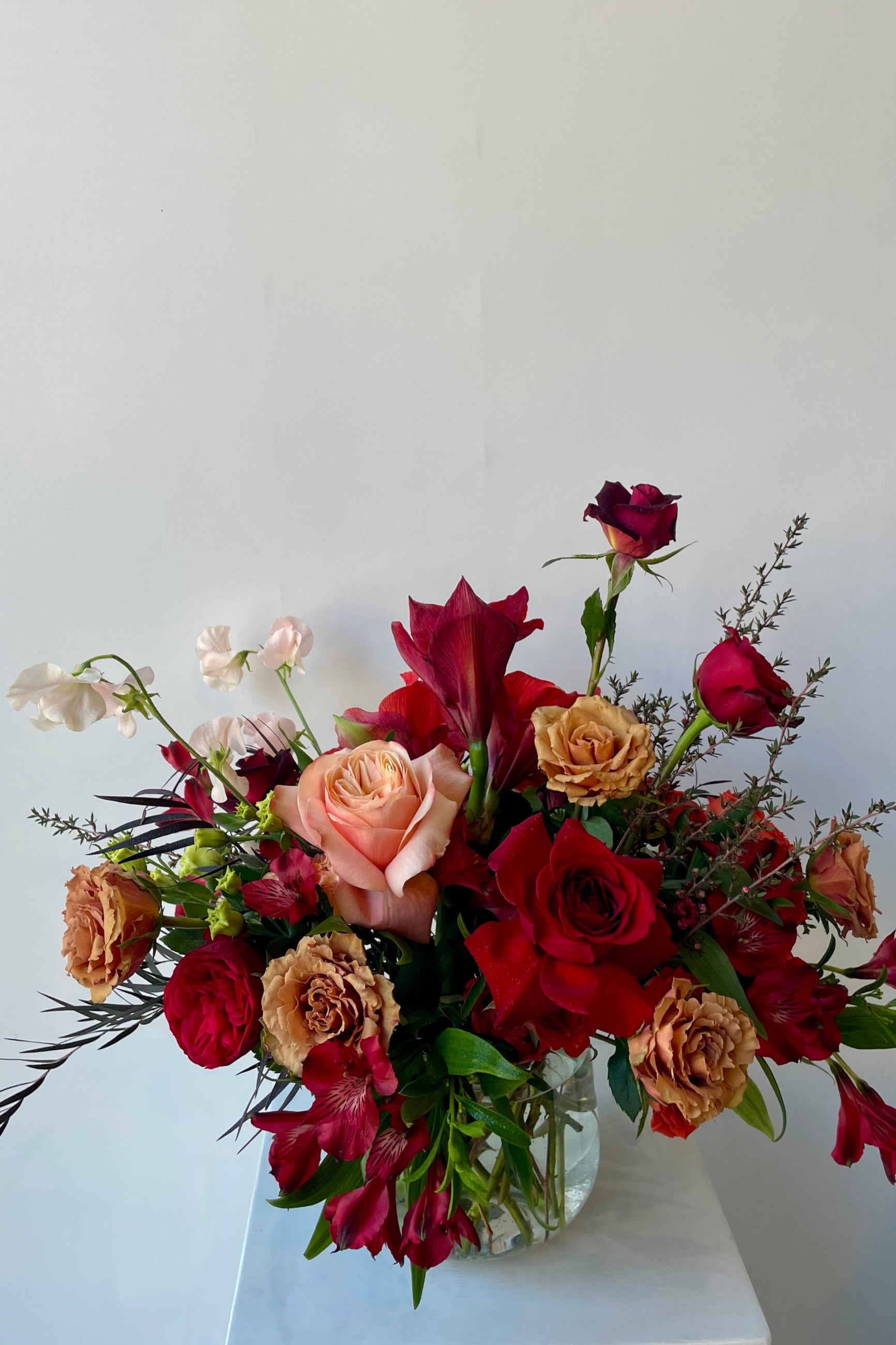 Earth floral arrangement at Sprout Home with Red and Toffee Roses and pink sweet pea styled in clear glass shown against a white wall. ©Sprout Home #size_$200