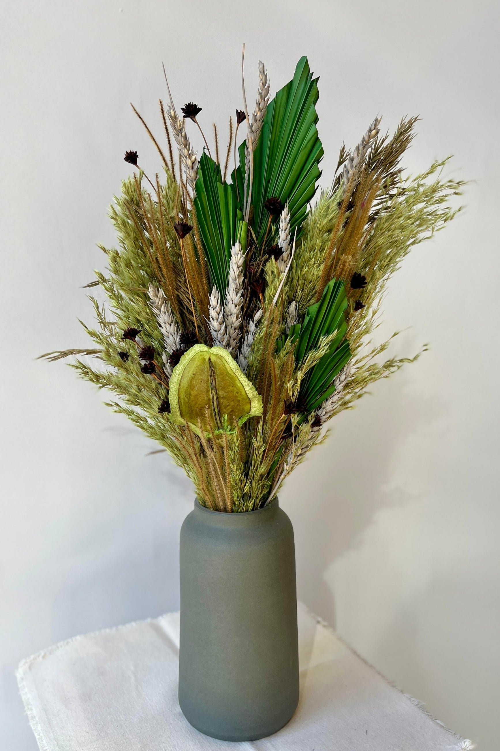 An arrangement of preserved floral and foliage with a variety of flower types in shades of green, brown, grey and gold against a grey wall. The arrangement is shown in a dark green vase. ©Sprout Home #size_$85