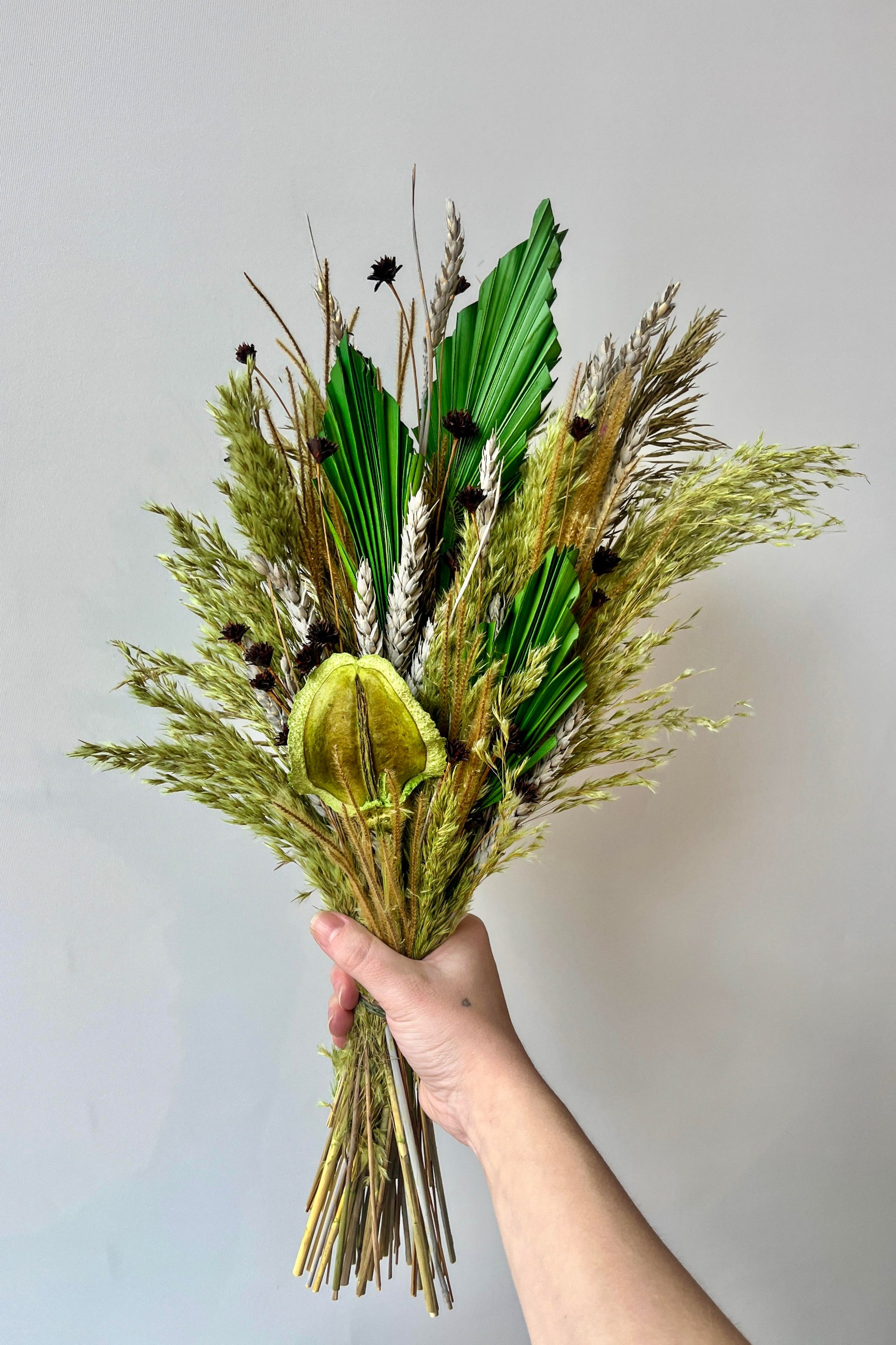 Hand holding preserved floral arrangement with a variety of flower types in shades of green, brown, grey and gold against a white wall. ©Sprout Home #size_$85