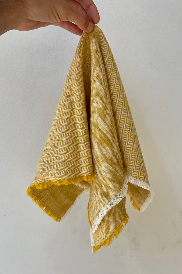 Photo of a Herrinbone pattern Mustard colored fringed napkin against a white wall.