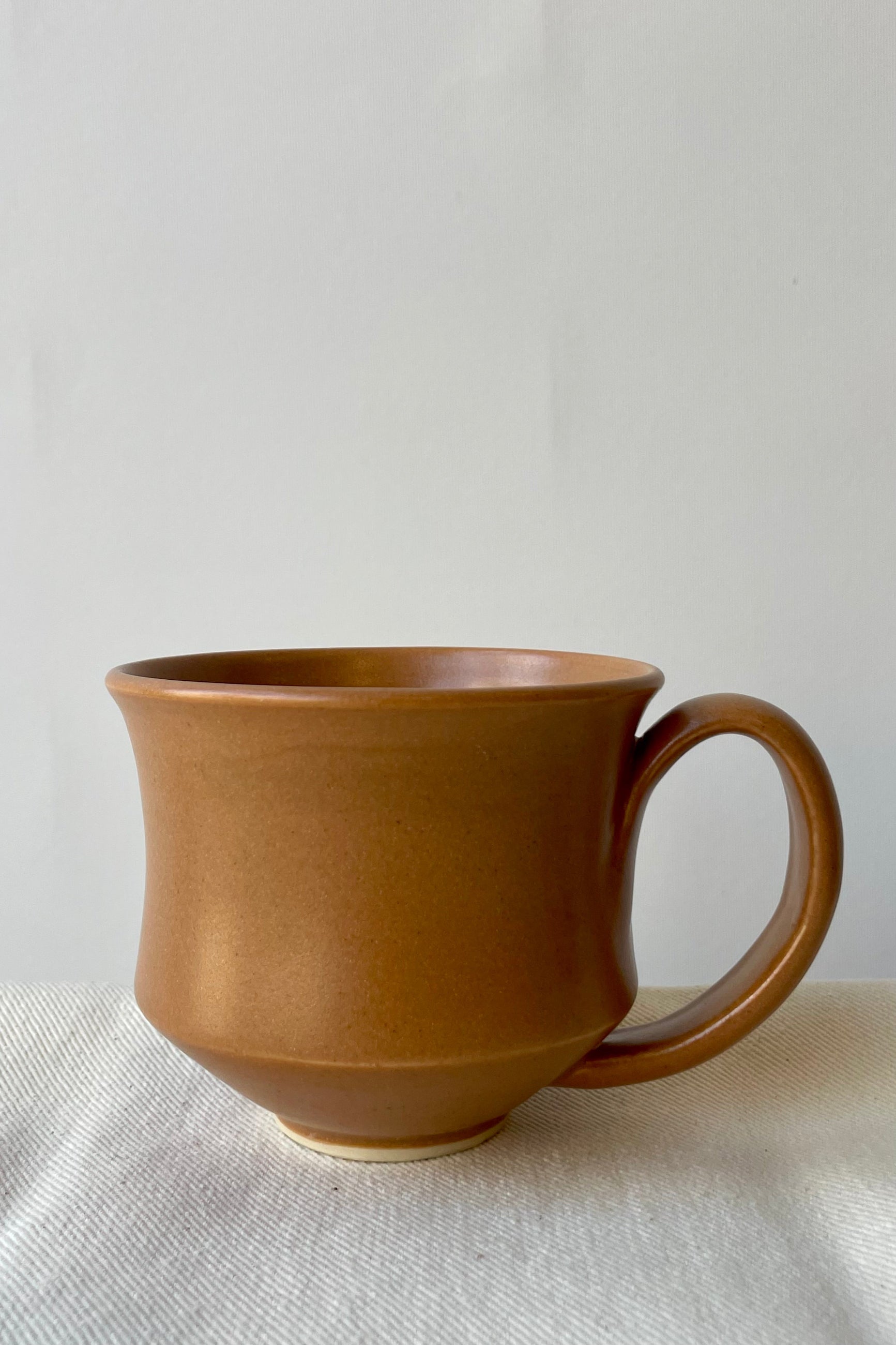 Photo of Little Fire Ceramics Houglass Mug with Carmel glaze. The mug sits on a white cloth against a white wall. ©Sprout Home
