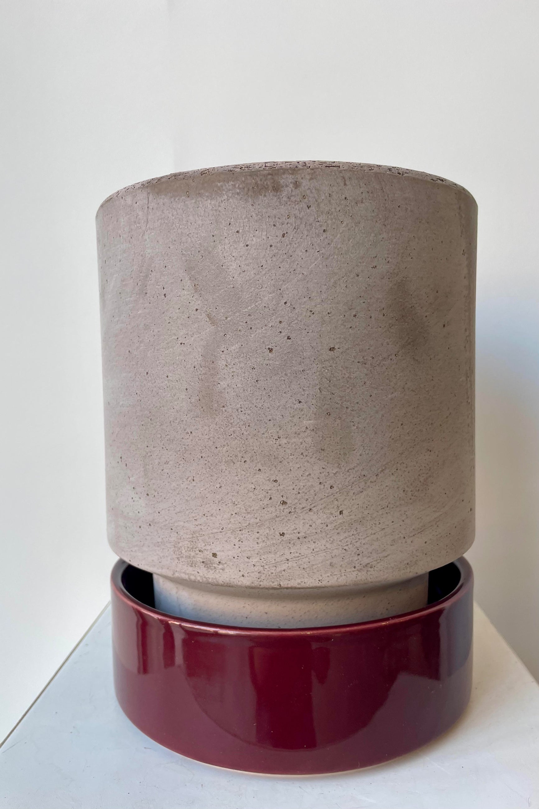 Photo of a Hoff Pot and saucer on a white table in a white room. The pot is gray and sits within to a deep burgundy glazed saucer. ©Sprout Home